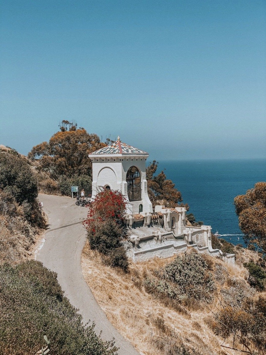A small white chapel with a checkered roof sits on a hillside overlooking the ocean, surrounded by dry vegetation and trees, with a dirt path leading up to it.
