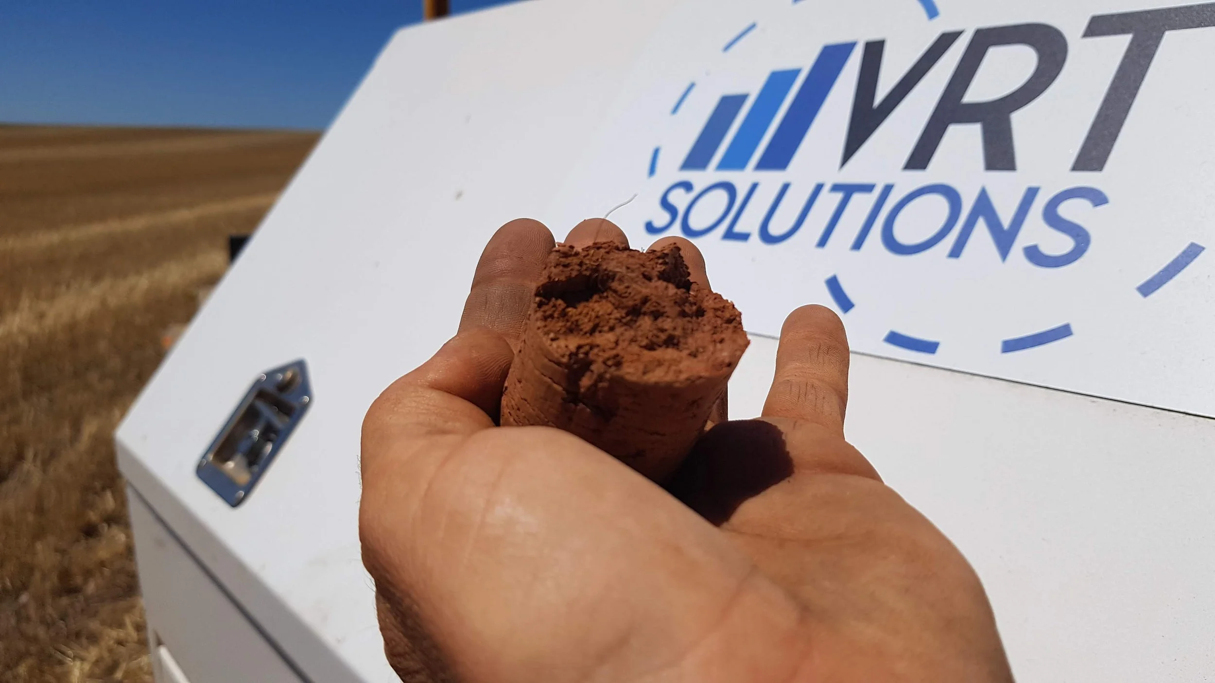 Person holding a piece of red rock or clay in front of a sign reading 'VRT Solutions' with a field in the background.