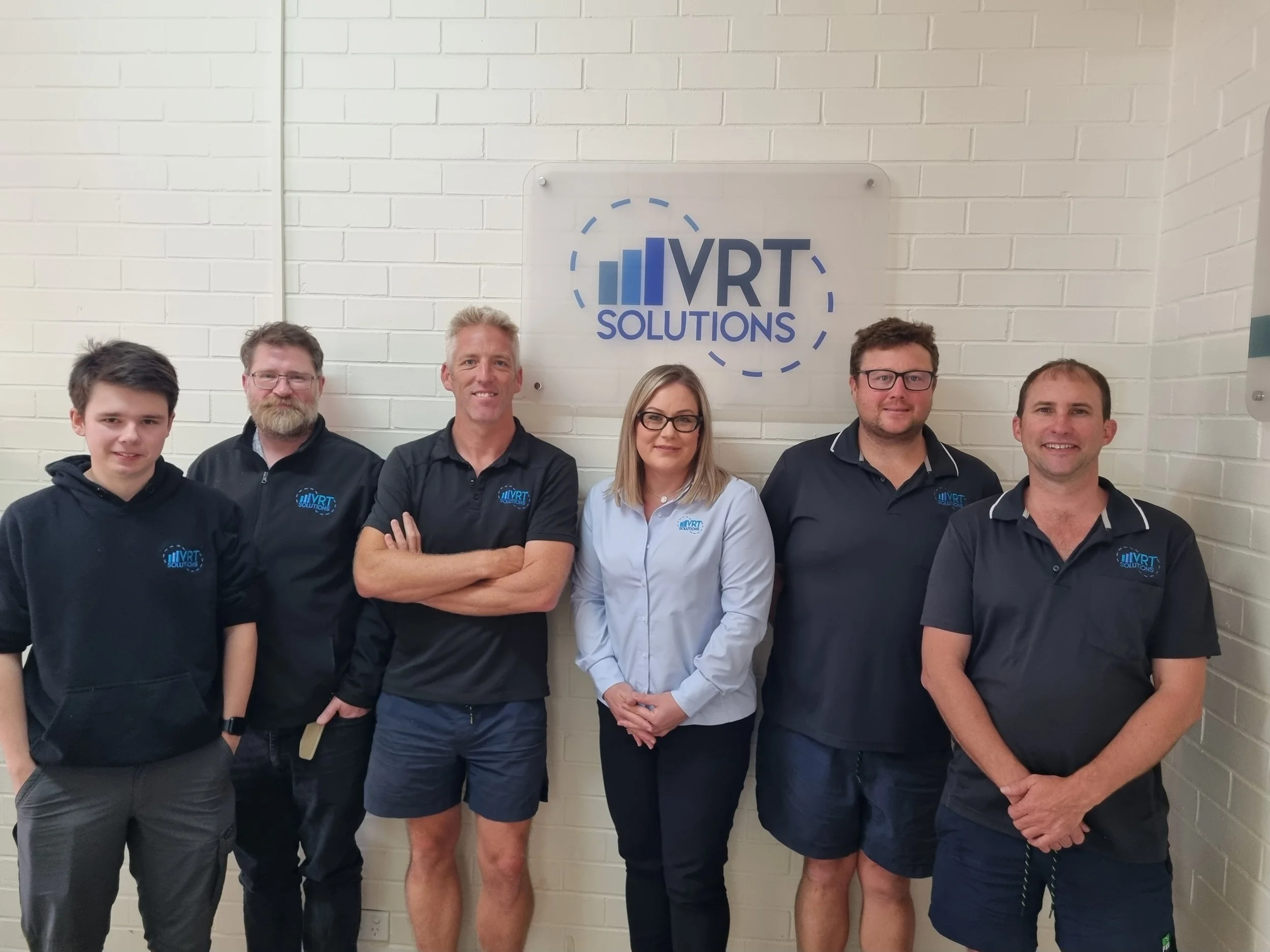 Group of six people standing in front of a wall with a sign that reads 'VRT Solutions'.
