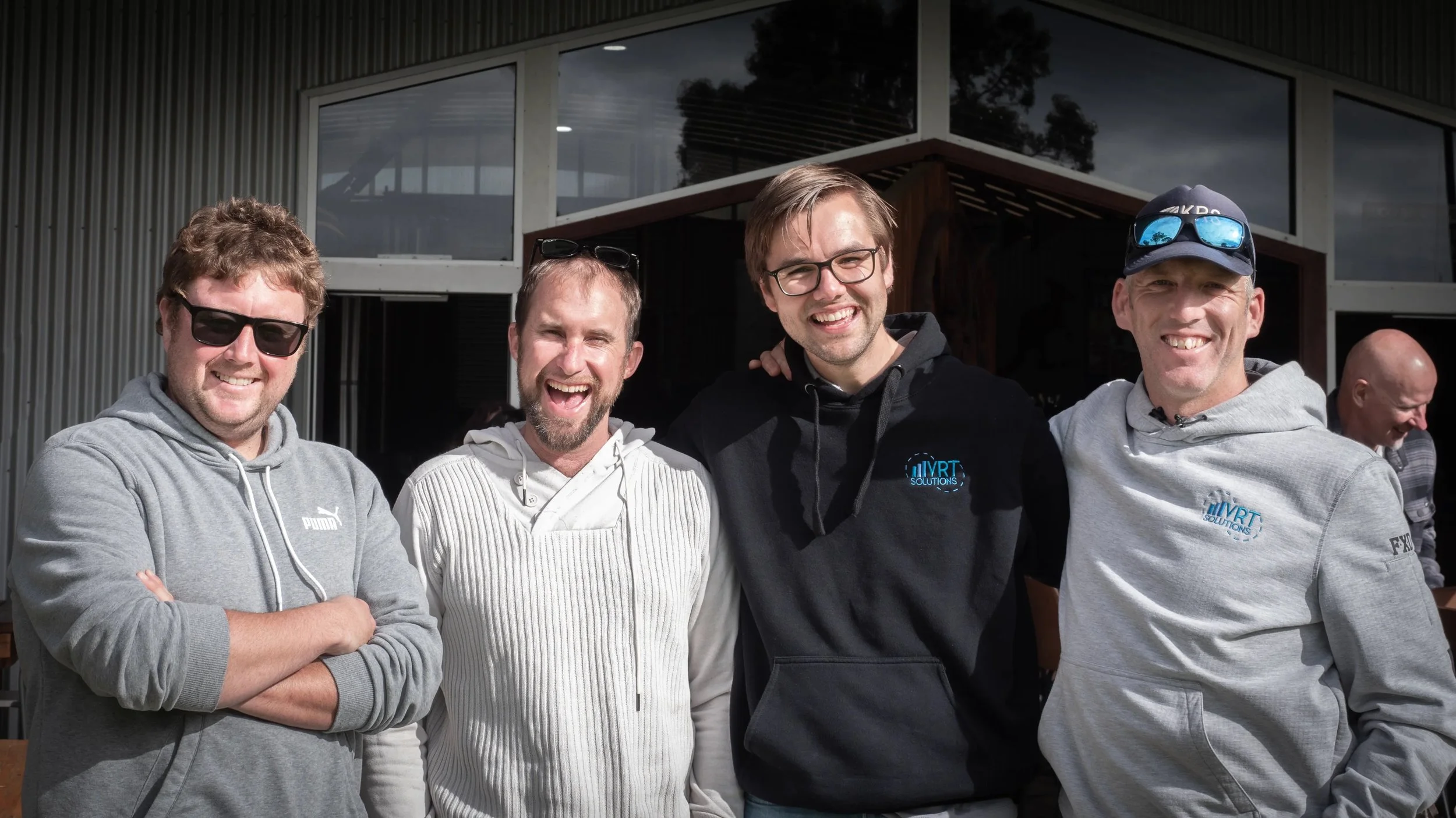 Group of four smiling men standing outdoors in front of a building with large windows, wearing casual and athletic clothing.