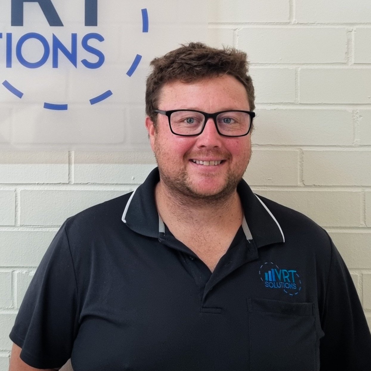 Man with glasses smiling, wearing a black polo shirt with a logo that says 'VRT Solutions,' standing against a white brick wall with a partial blue text sign.