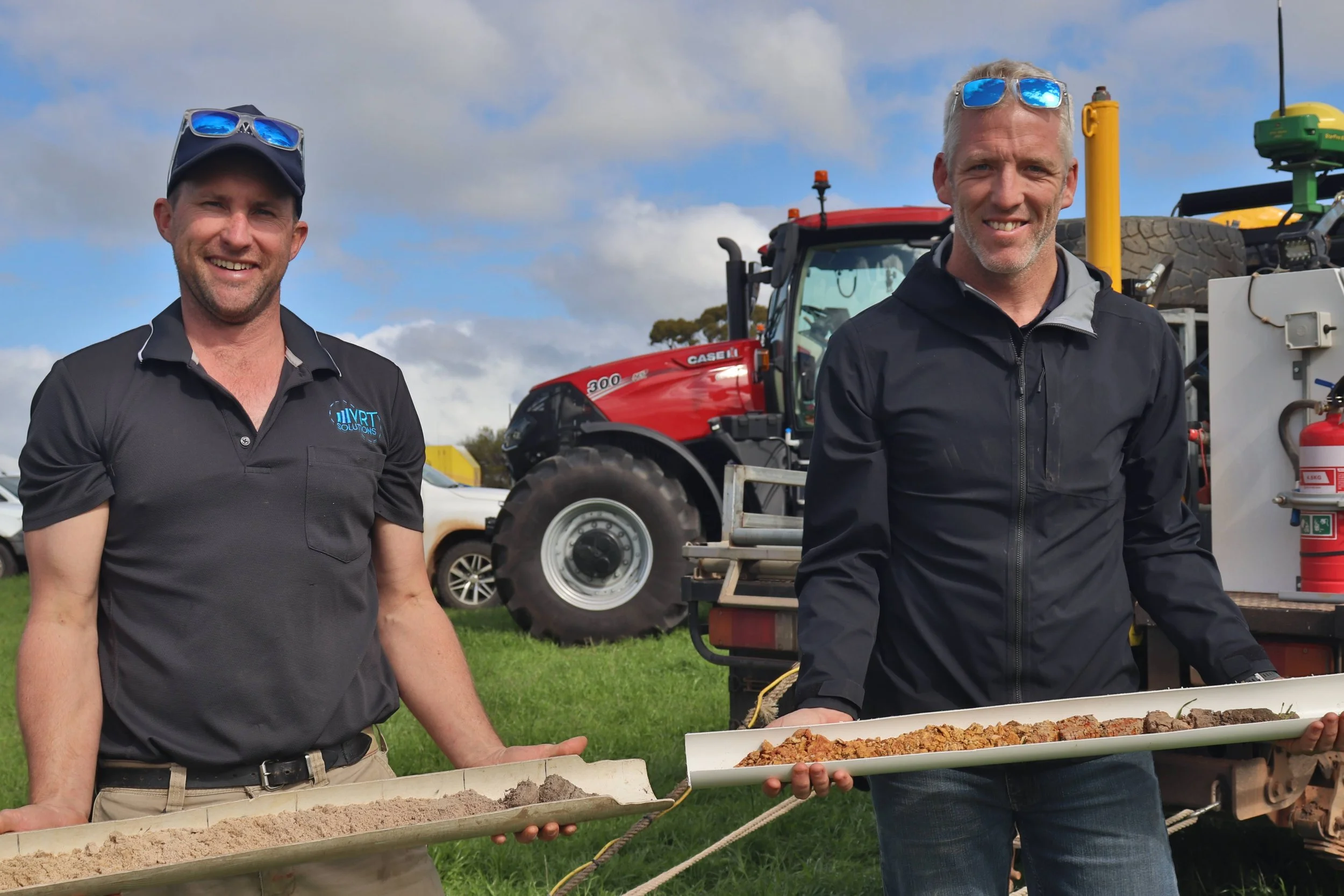 Two men standing outdoors, each holding a long tray with food, with farm equipment and a red tractor in the background.