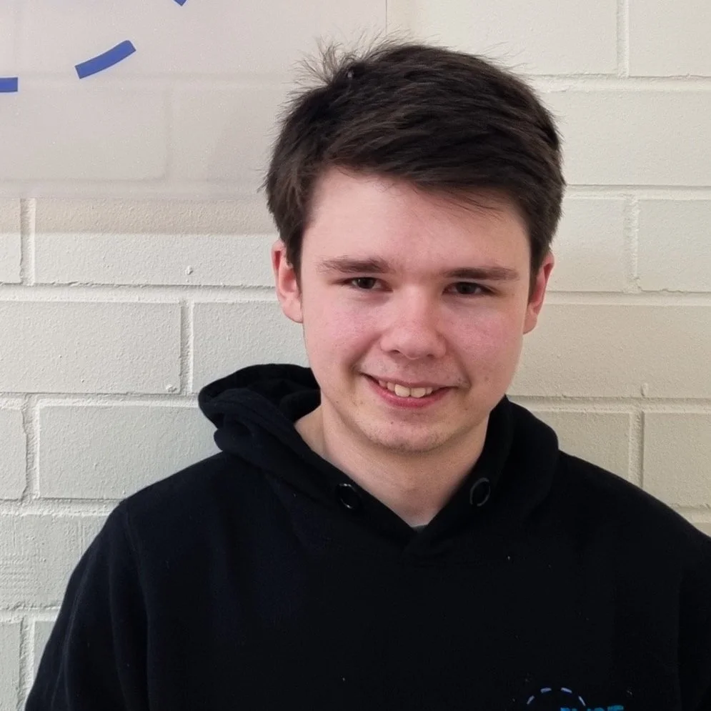 A young man with short brown hair smiling, wearing a black hoodie, standing in front of a cream-colored brick wall.