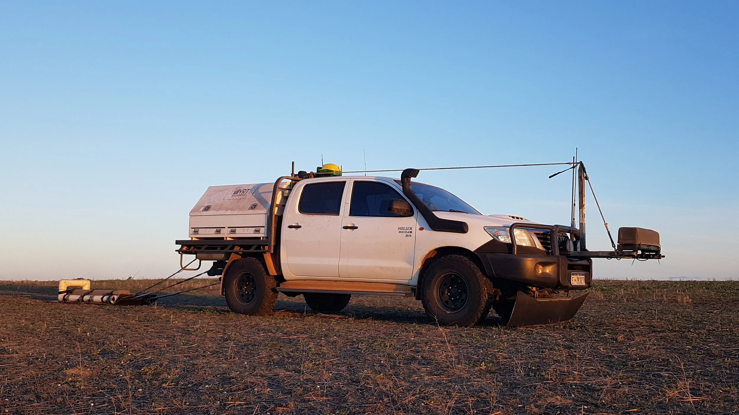 A white Toyota ute equipped with scientific or technical equipment, including a large white container in the truck bed, an antenna, and other apparatus mounted on the front and sides, is parked in a flat, open field during sunset.