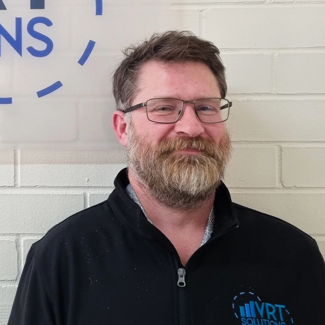 A man with glasses and a beard standing against a white brick wall, wearing a black jacket with a blue VRT Solutions logo.