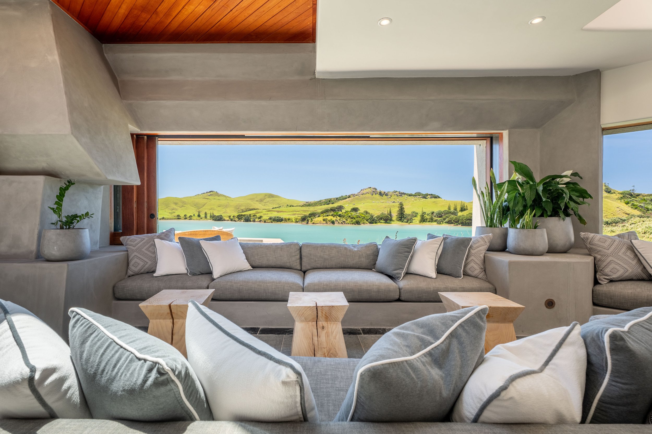 Living room with large window view of green hills and water, gray sofas, white and gray pillows, and potted plants.