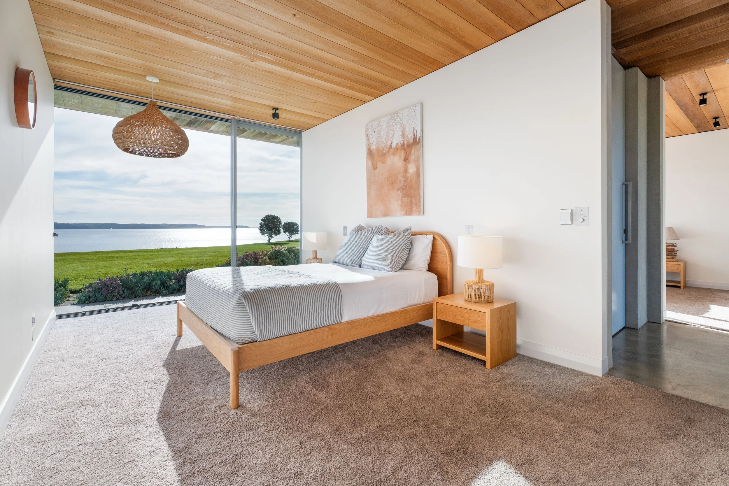 Modern bedroom with a large window overlooking a lake and greenery, wooden ceiling, minimal decor, wooden bed with striped bedding, two bedside lamps, a piece of abstract art on the wall.