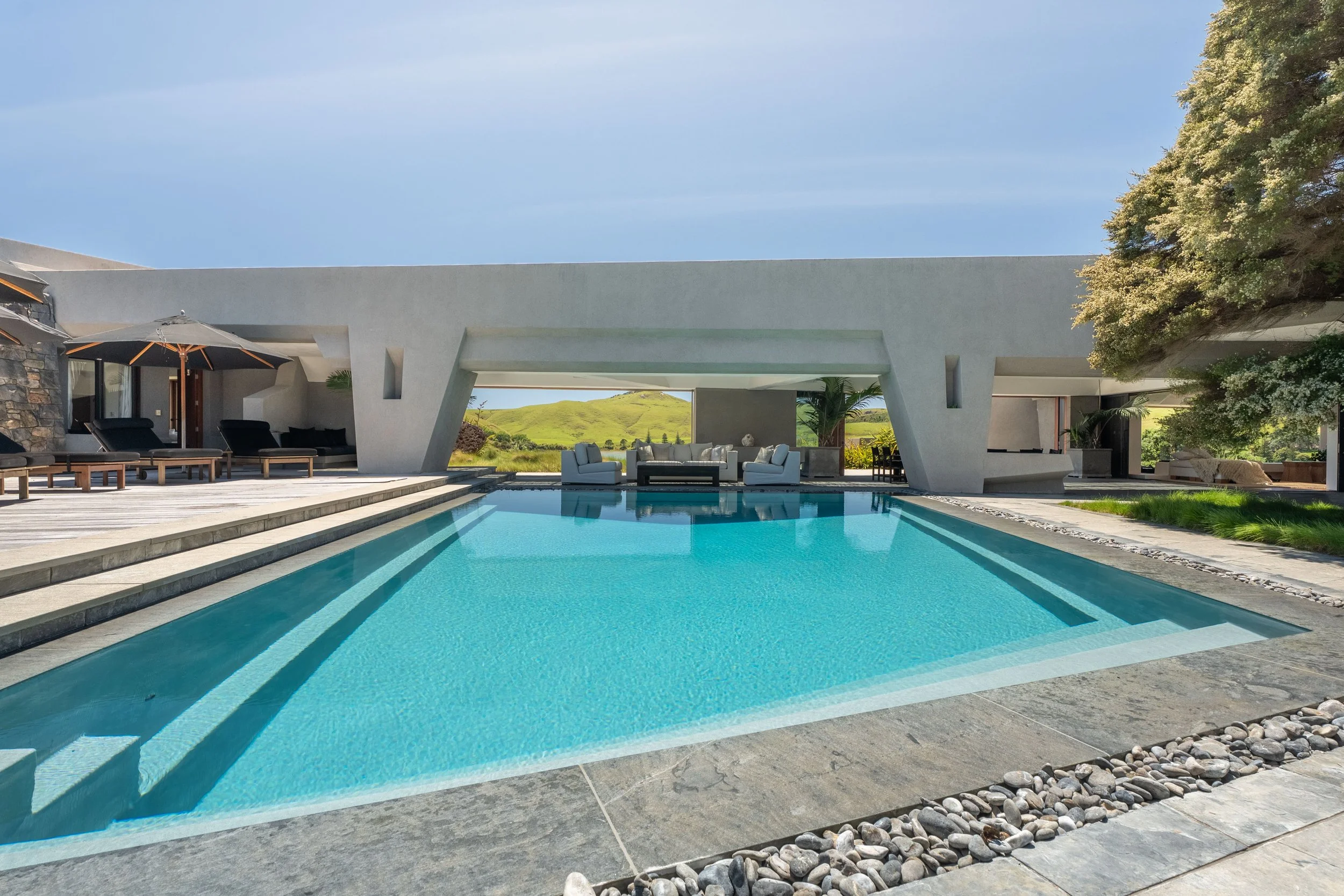 Modern house with a swimming pool in front, outdoor lounge chairs, umbrellas, and lush green landscape with hills in the background.