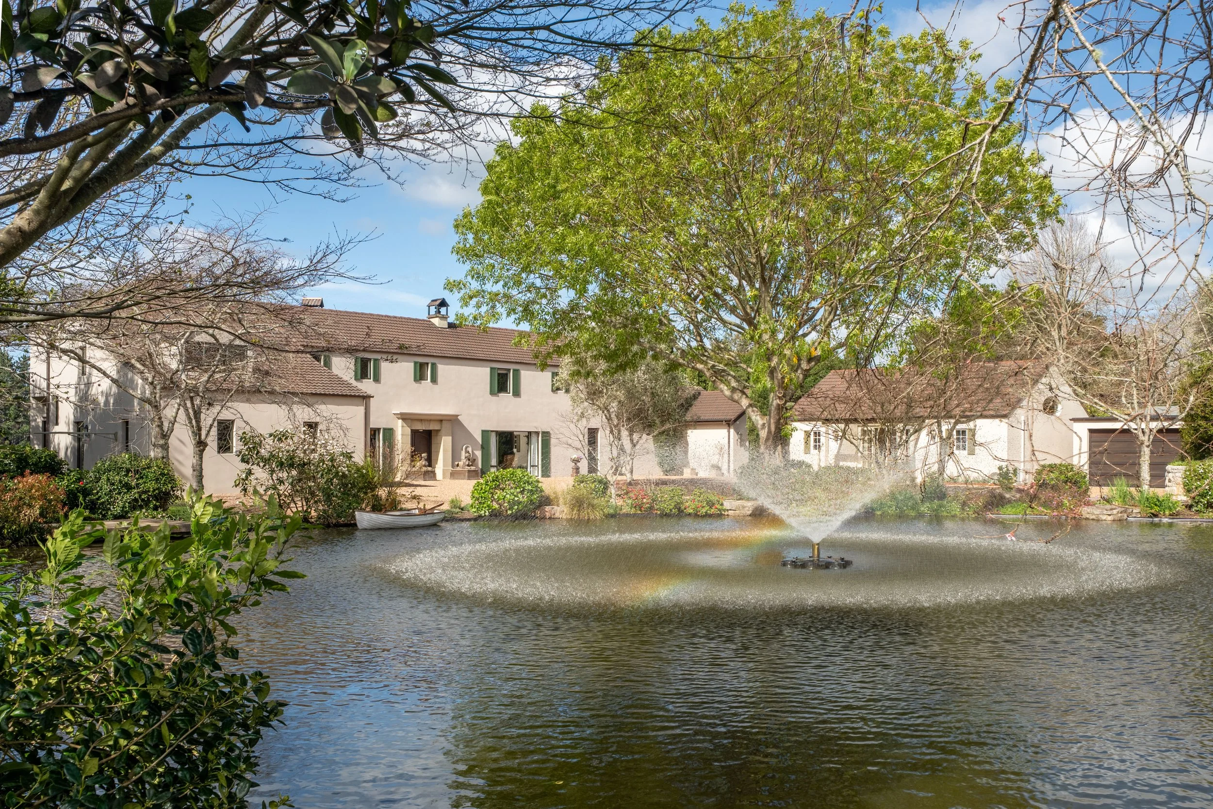 A pond with a fountain amidst lush bushes and trees, with a residential house and garage in the background under a partly cloudy sky.