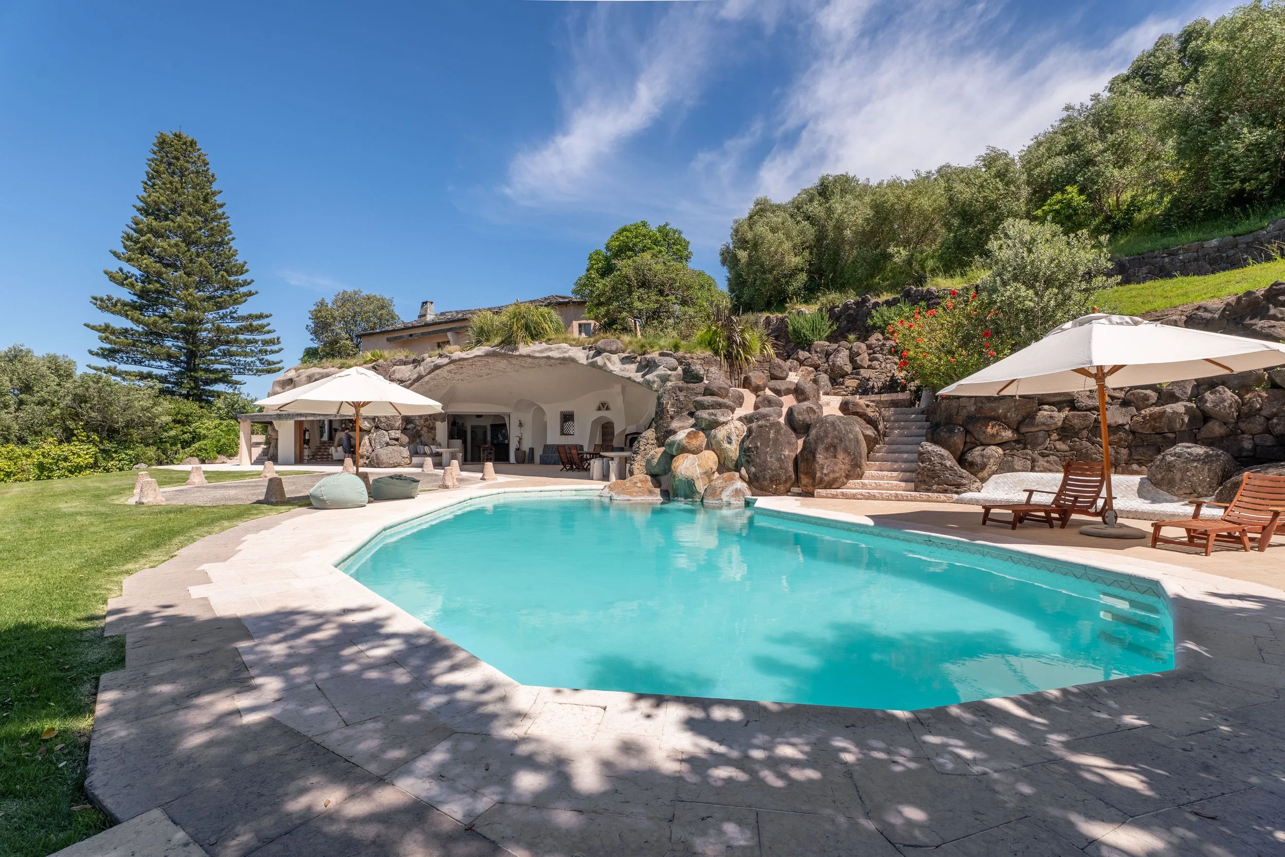 Swimming pool in a backyard with umbrellas, lounge chairs, trees, rocks, and a house built into a hillside under a blue sky.