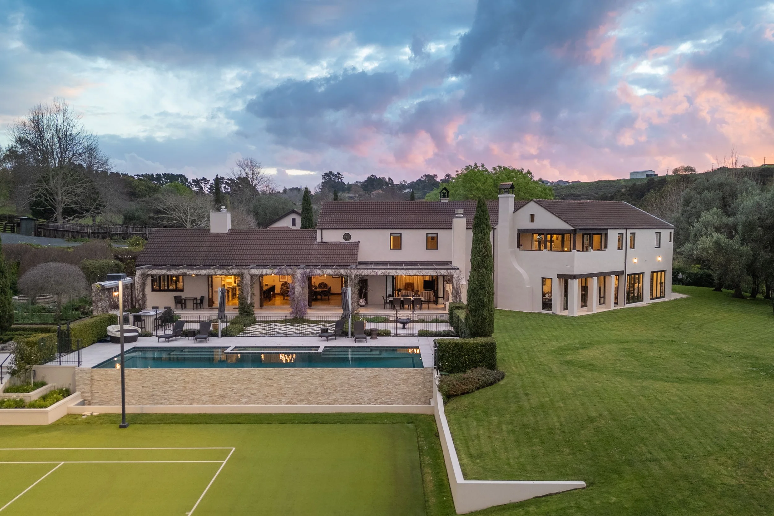 Large modern house with multiple levels, white exterior walls, large windows, and a patio area. There is a swimming pool, a tennis court, and well-maintained green lawns surrounding the house. The sky shows a colorful sunset with clouds.