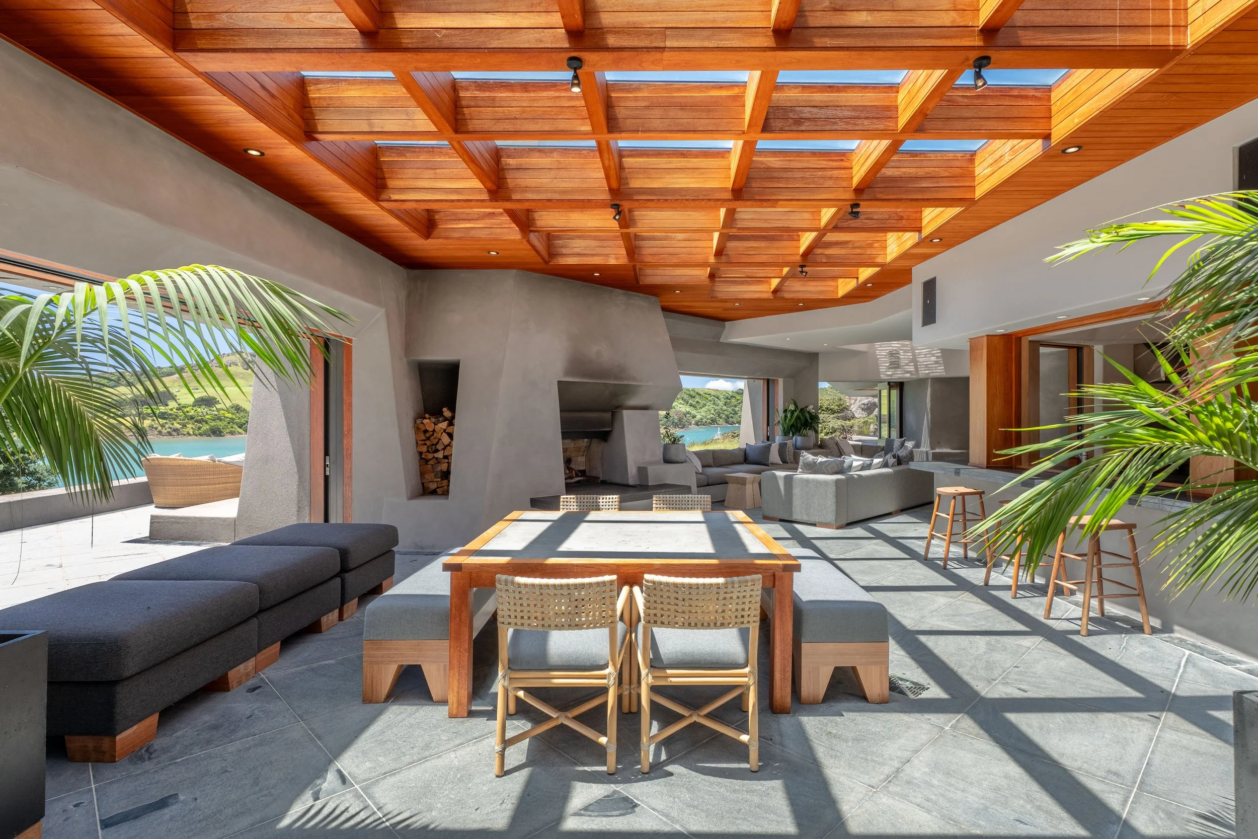 Spacious modern living room with large windows, wooden ceiling, gray sofa, dining table with benches, and outdoor view of landscape and water