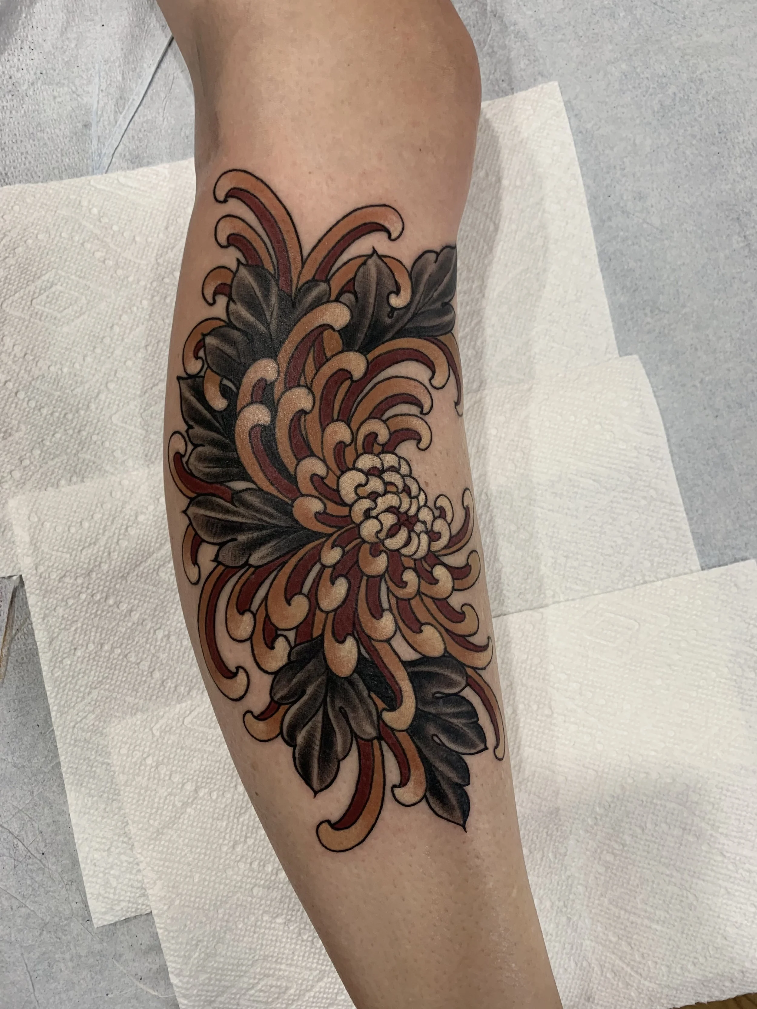 A colorful floral tattoo on a person's arm featuring a large flower with black leaves and swirling petals in shades of red, orange, and beige.