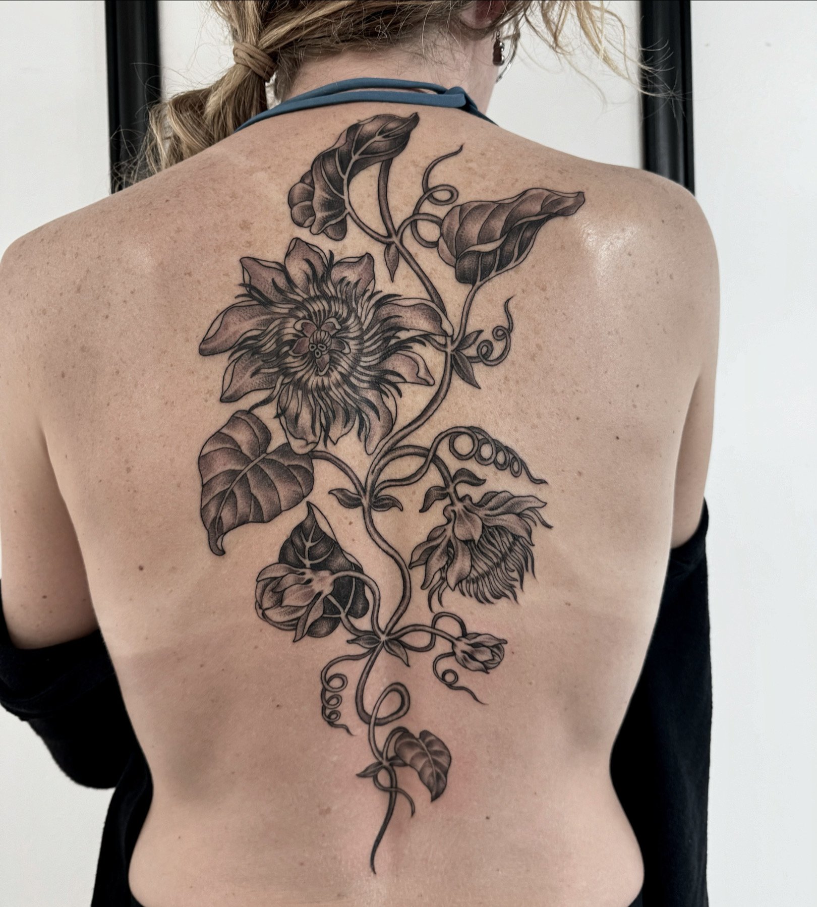 Back, woman with a large black and gray floral tattoo extending from upper back to lower back, featuring sunflowers and leaves.