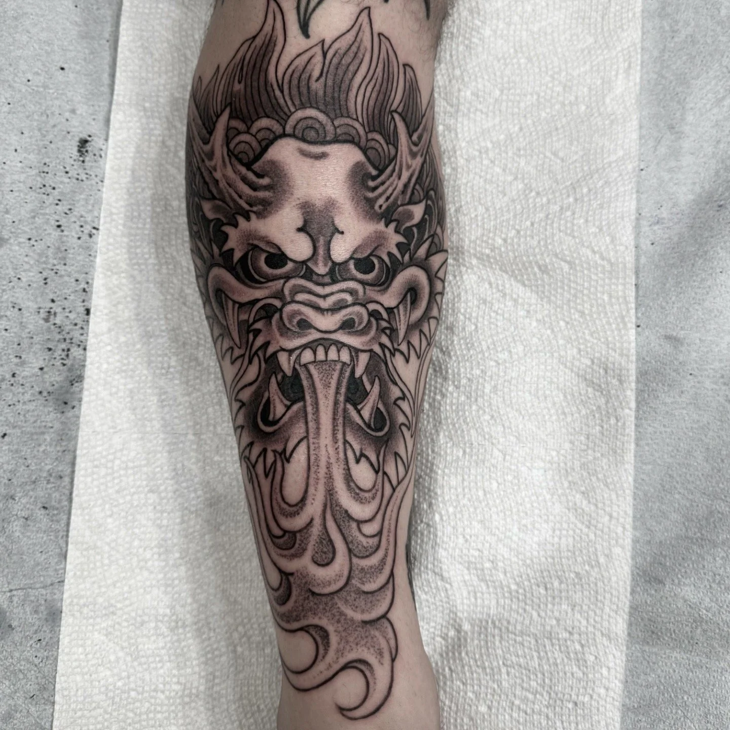 Black and gray tattoo of a fierce demon or mythical creature with large eyes, sharp teeth, a protruding tongue, and intricate detailing on the face and head, covering the forearm.