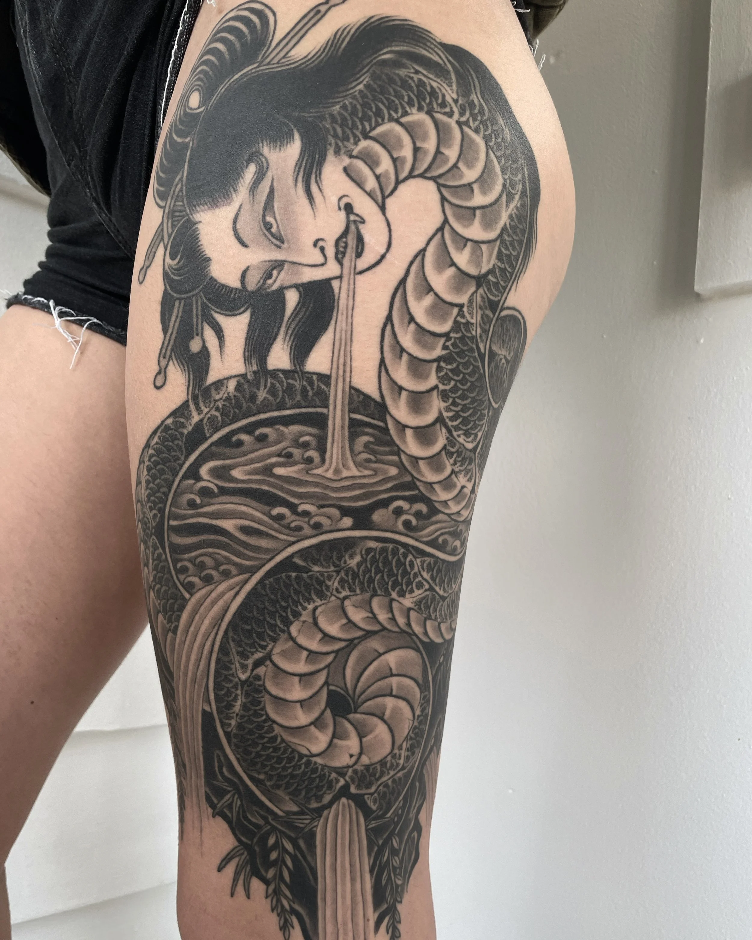 Black and gray tattoo on a person's upper thigh depicting a woman with traditional Japanese hairstyle and makeup, biting a snake which curls around her and extends downward, with flowing water and waves in the background.
