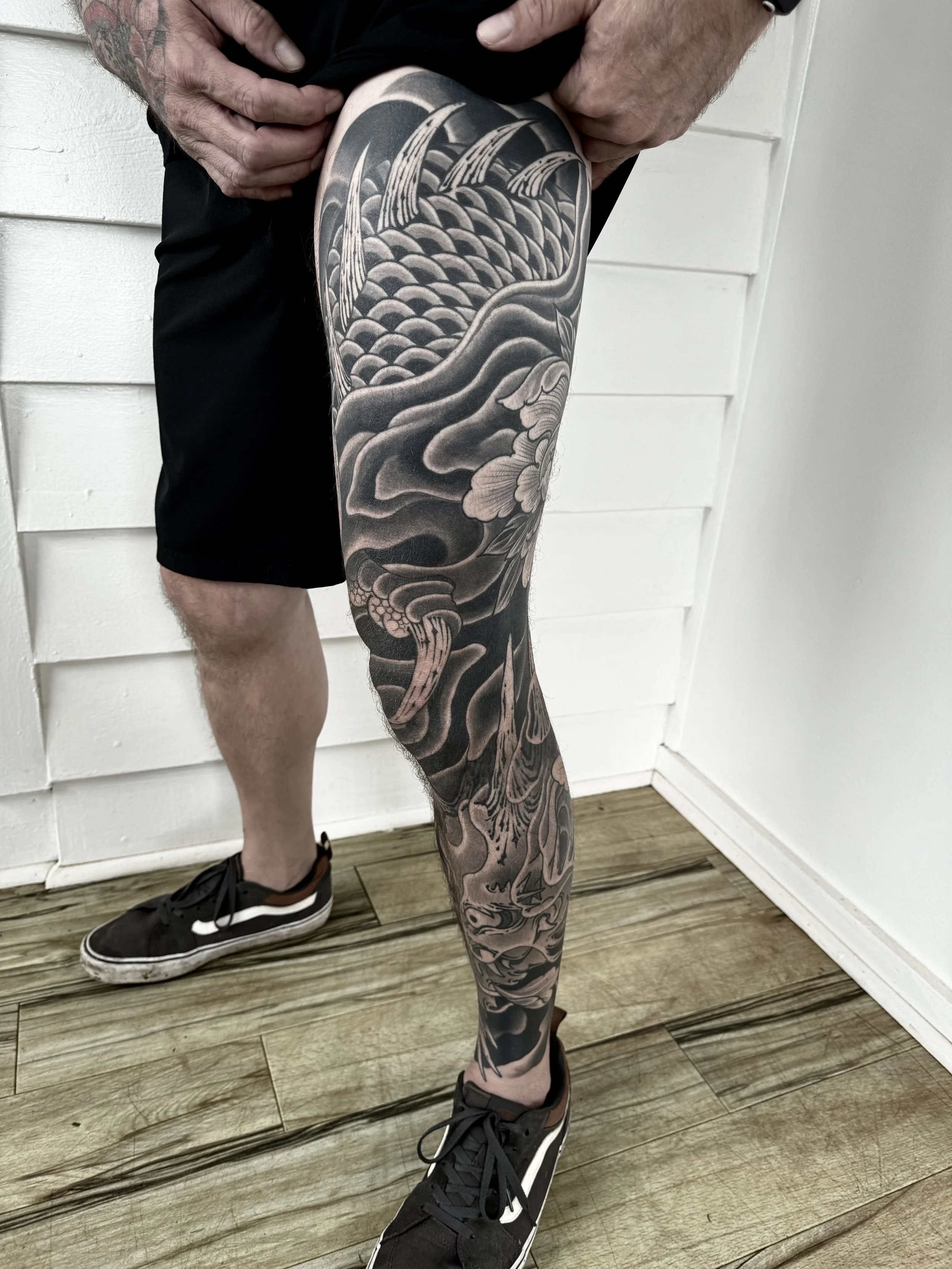 Close-up of a person's leg with a detailed black and gray Japanese-style tattoo featuring a dragon, clouds, and floral elements.