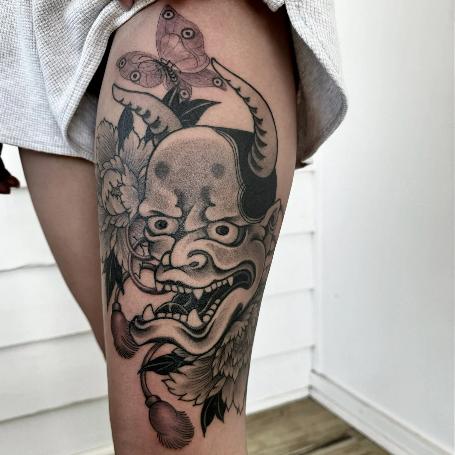 Black and gray tattoo of a fierce mythical creature with a lion-like face, horns, and floral details on a person's thigh.