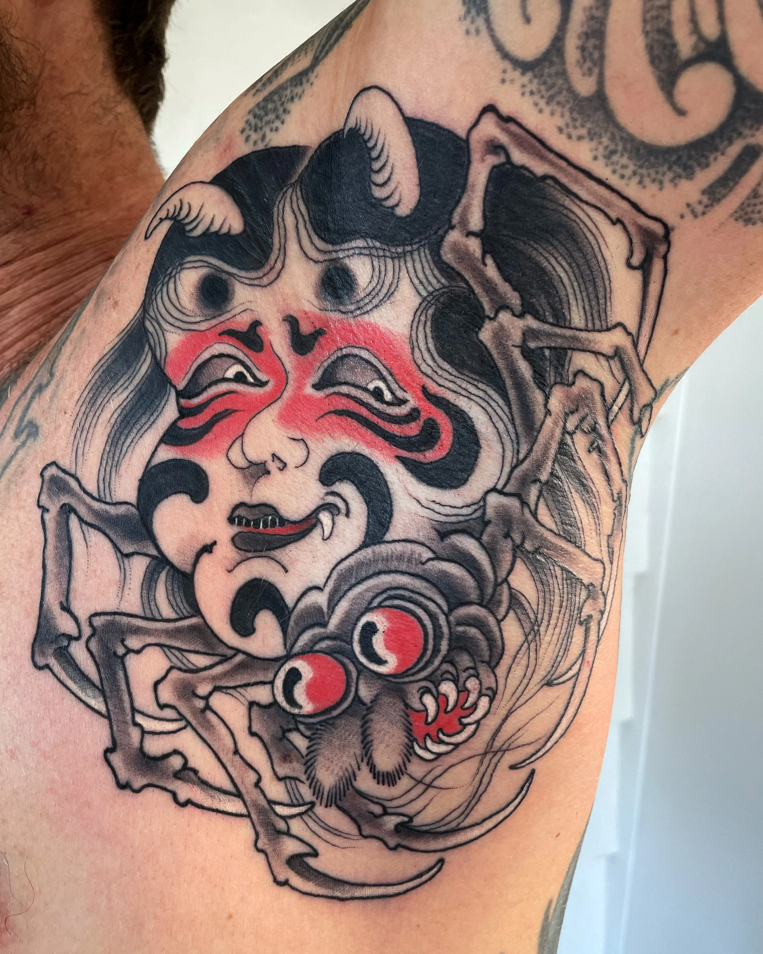 Tattoo of a traditional-style Japanese demon mask, with red and black details, and a spider with red eyes and sharp fangs underneath.