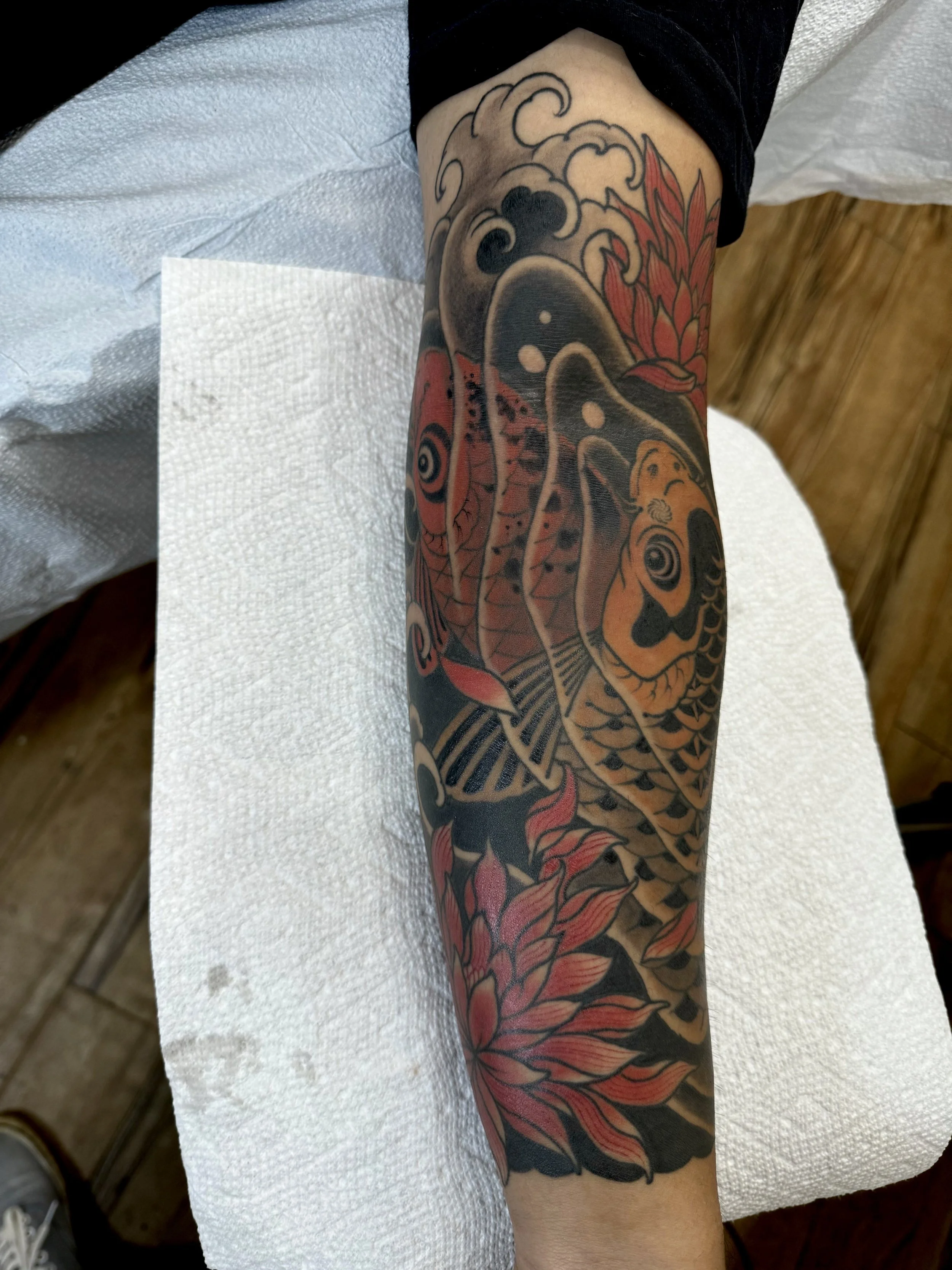 A tattoo sleeve featuring a koi fish, red and black flowers, and waves on a person's arm.