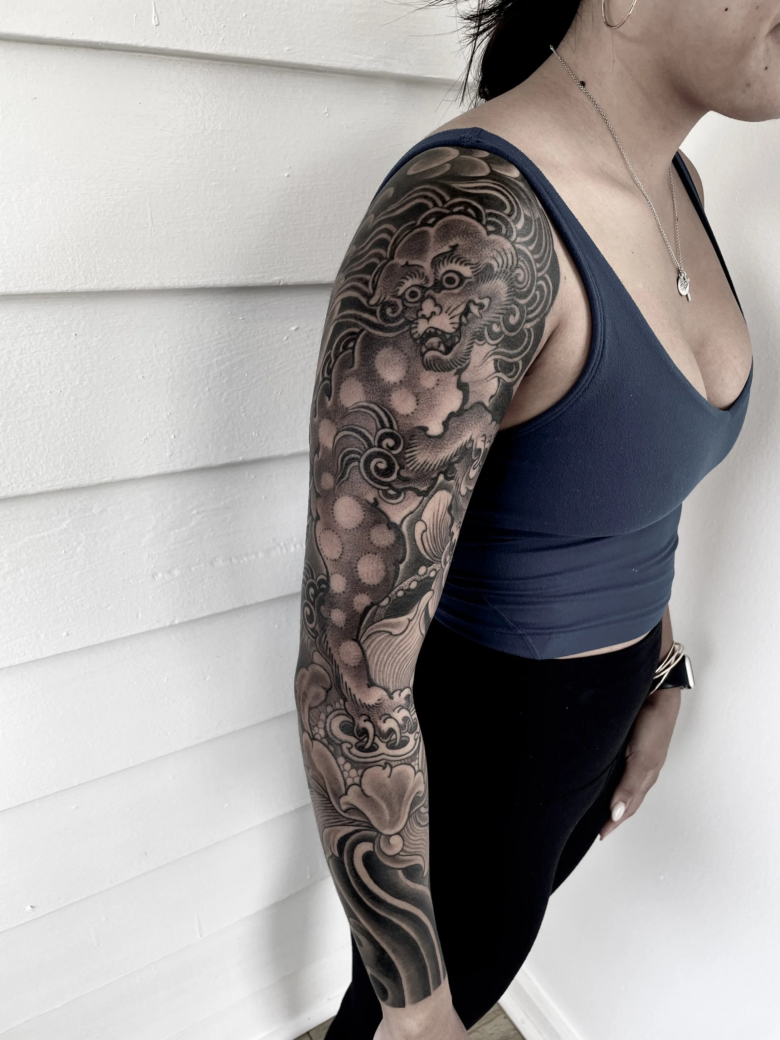 A woman with a detailed black tattoo sleeve of a lion on her upper arm, wearing a dark blue sleeveless top, standing against a white wall.