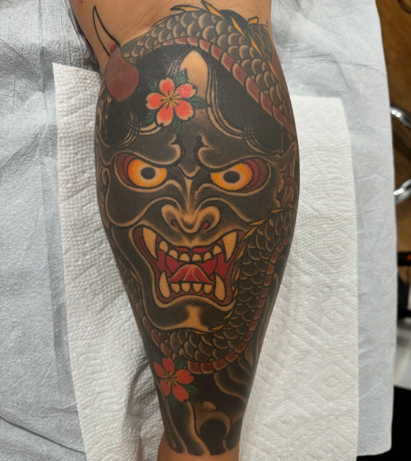 Tattoo of a fierce mythical creature with a dragon-like face, orange eyes, sharp teeth, surrounded by red flowers and a snake, on a person's leg.