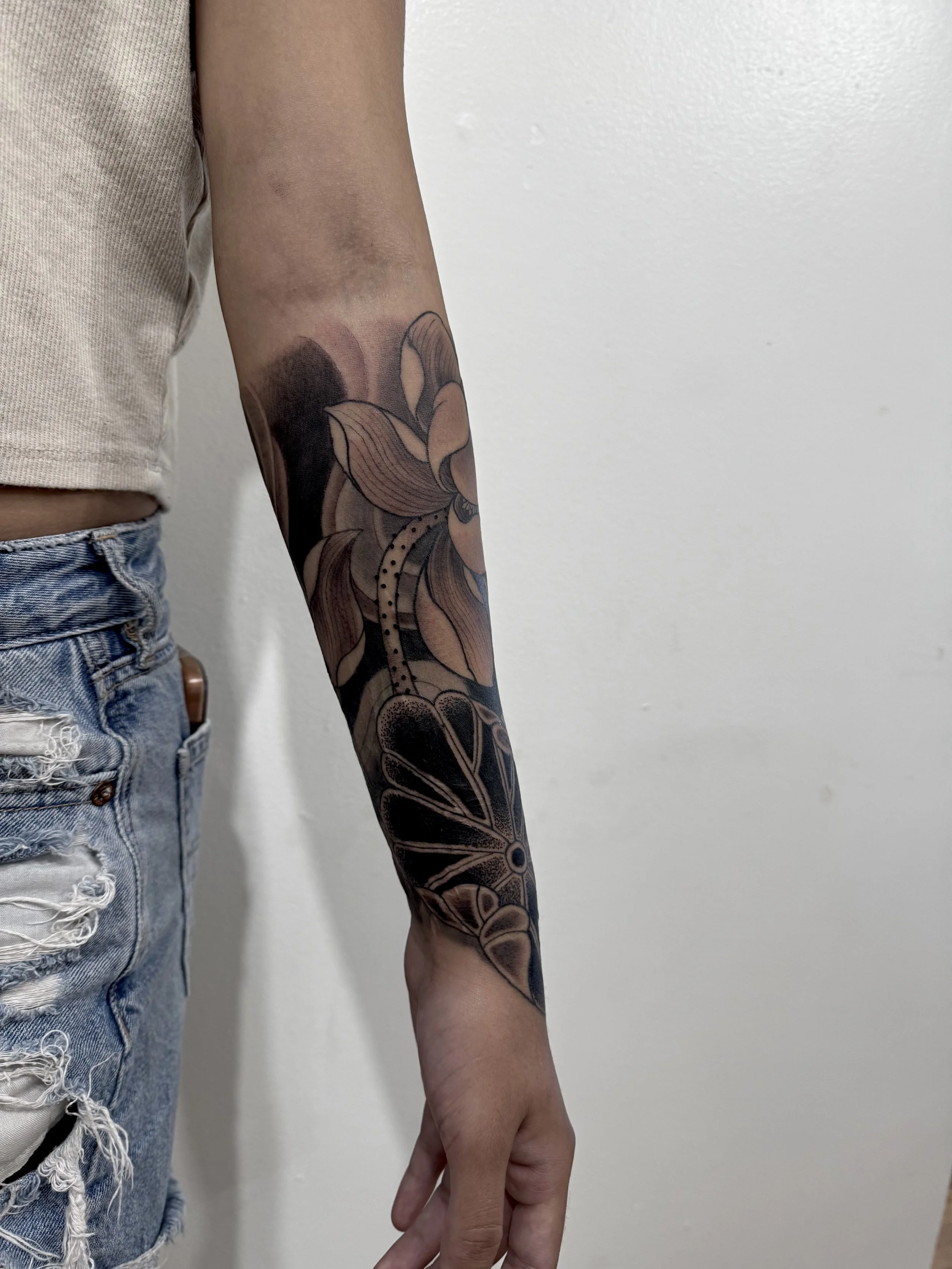 A person's arm with a black and beige floral tattoo extending from the wrist to the elbow, visible part of a beige shirt and distressed blue jeans.