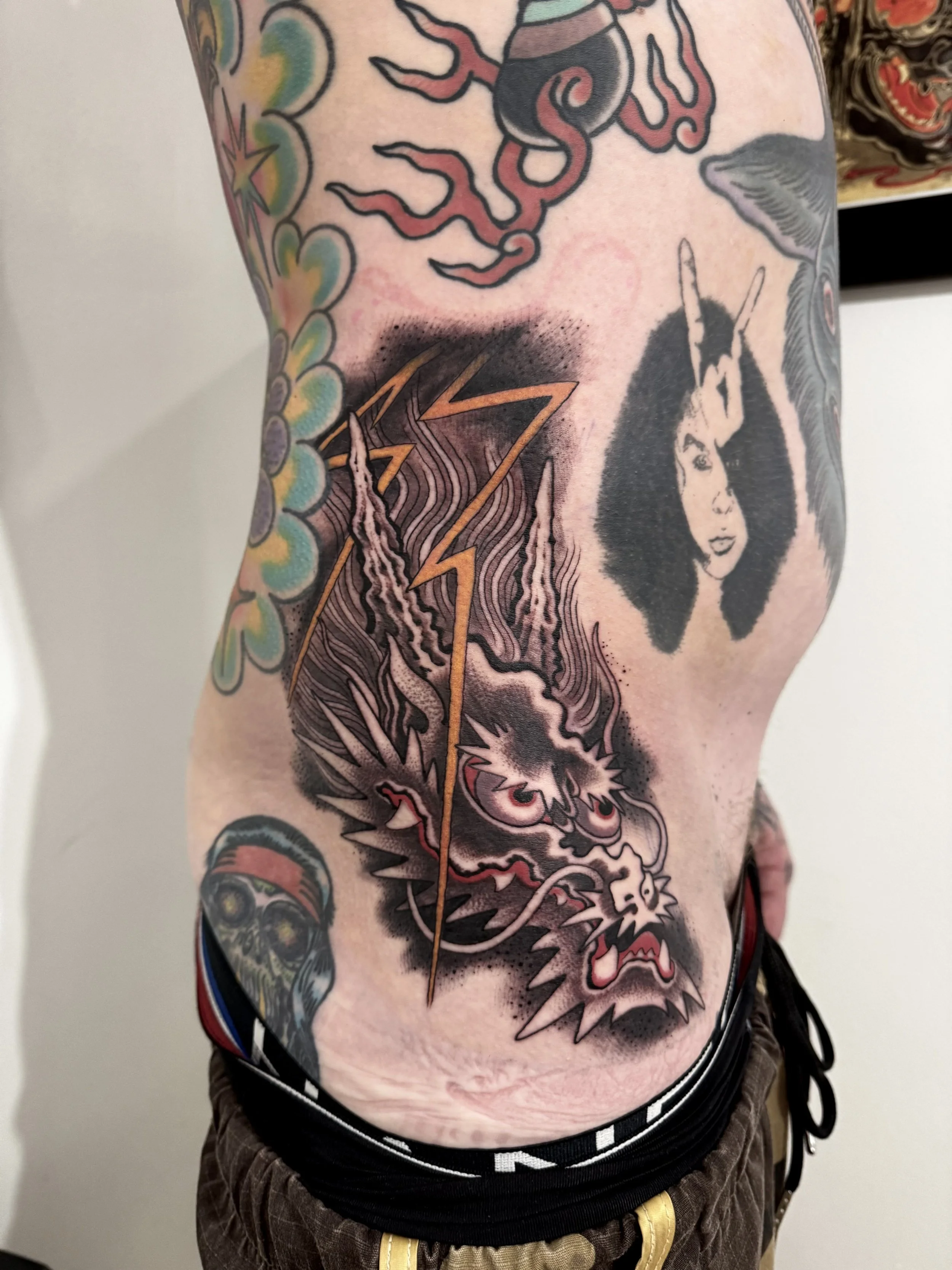 Tattoo sleeve on a person's torso, featuring a black and white portrait of a woman with curly hair and a unicorn horn, a colorful wolf head, a lightning bolt, and a skull with eye patches.