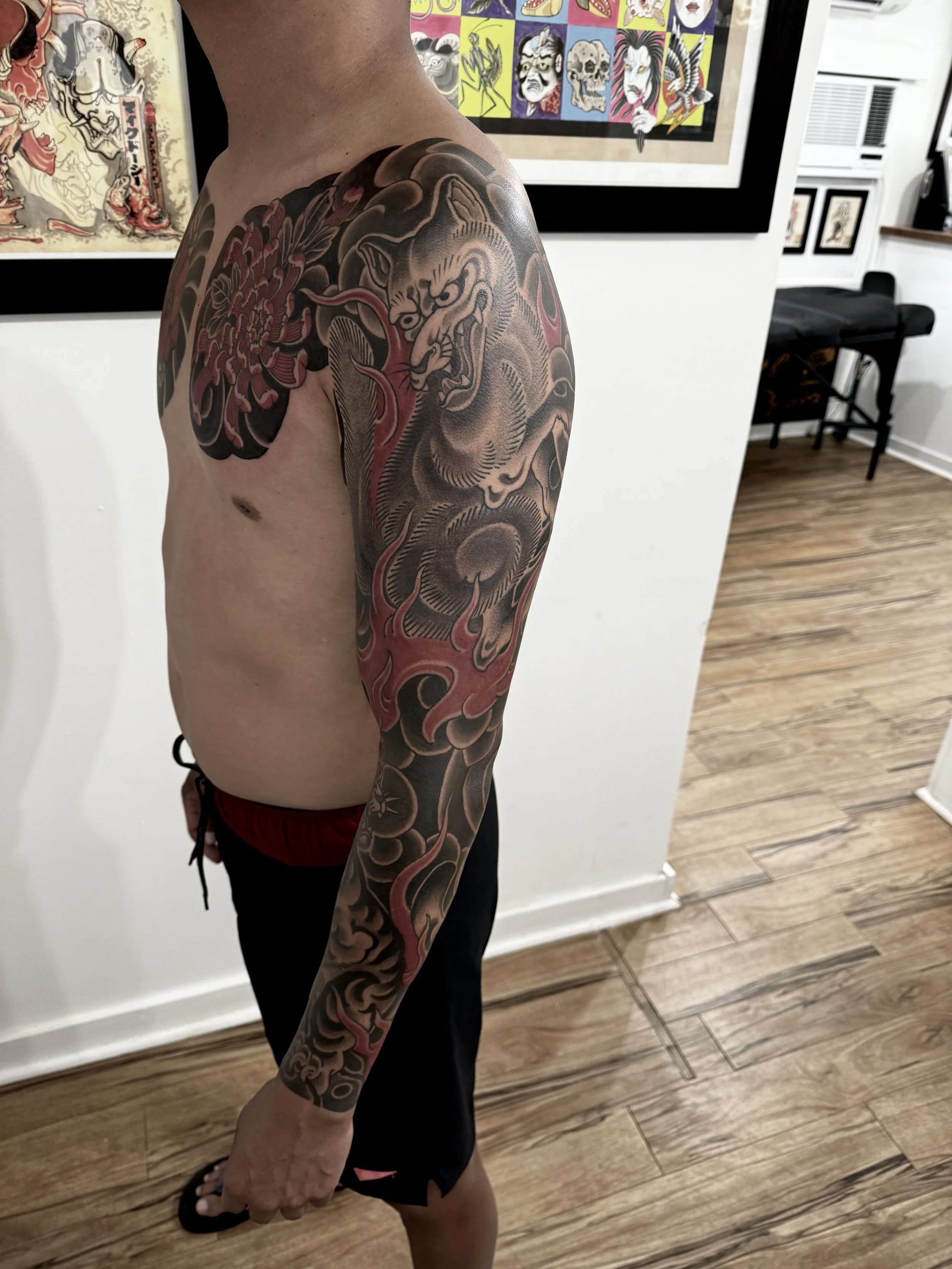Tattooed man with a large dragon tattoo on his upper arm, shoulder, and chest, standing indoors in front of artwork and framed pictures.