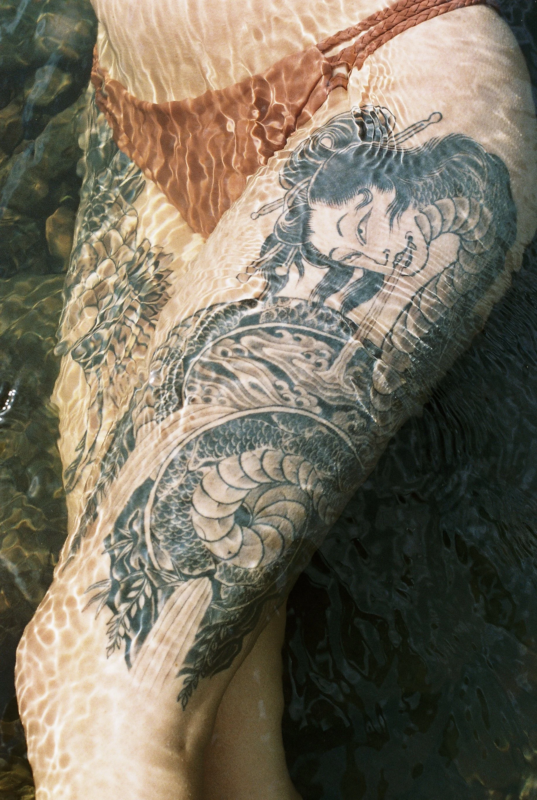 A person's arm submerged in water with a detailed black tattoo of a woman with flowing hair and a dragon tattooed around the arm.