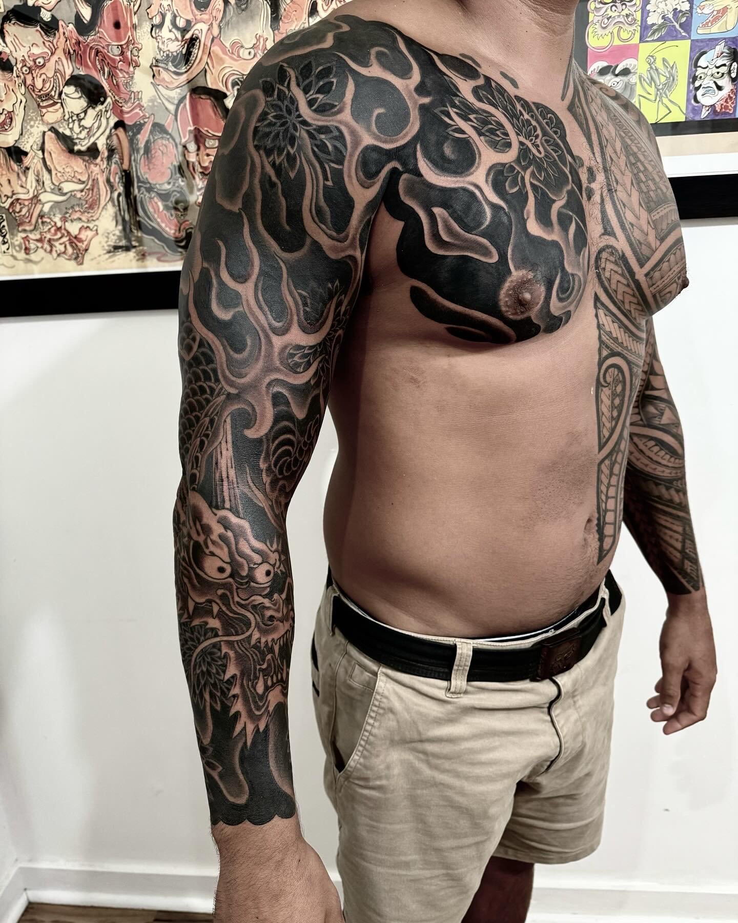 A man with extensive black ink tattoos covering his upper arm, shoulder, chest, and part of his side. The tattoos include intricate patterns and figures, with some areas filled with solid black and others featuring detailed line work. He is shirtless