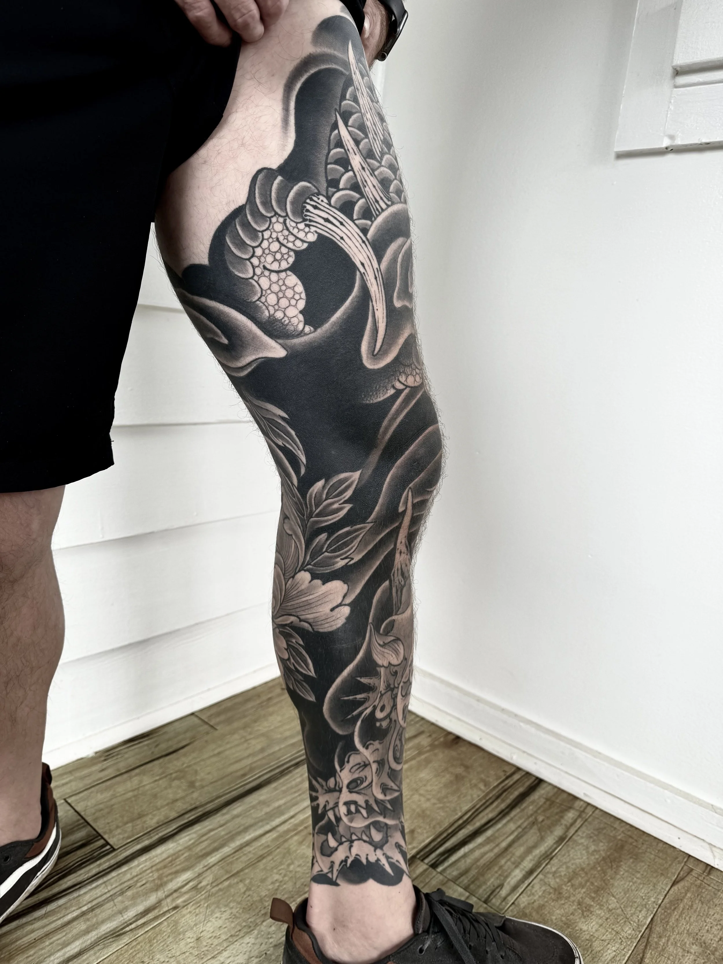 A person's leg with a large black and gray tattoo featuring a dragon and floral elements.