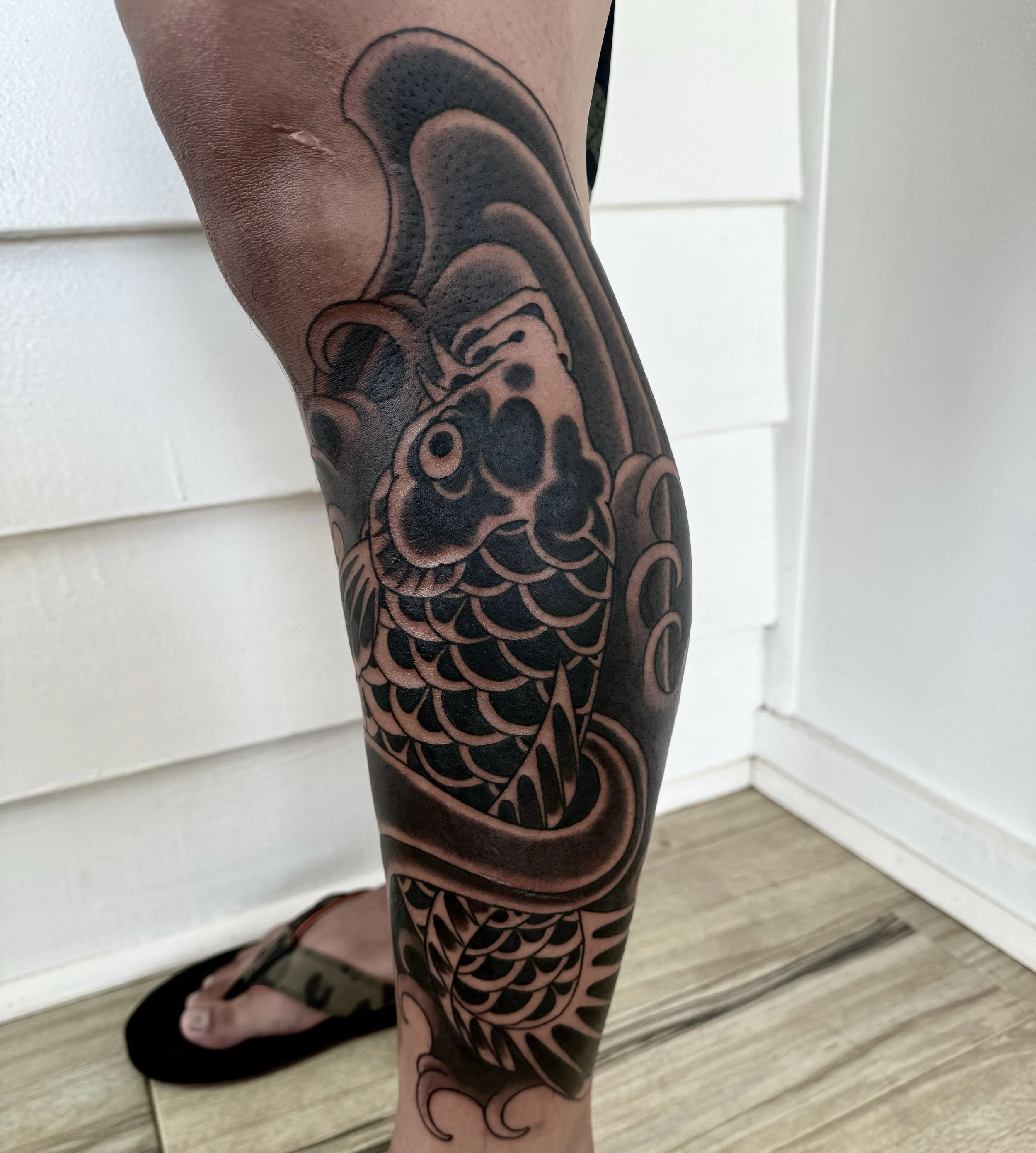 Tattoo of a koi fish with detailed scales on a person's leg, with dark shading and bold outlines.