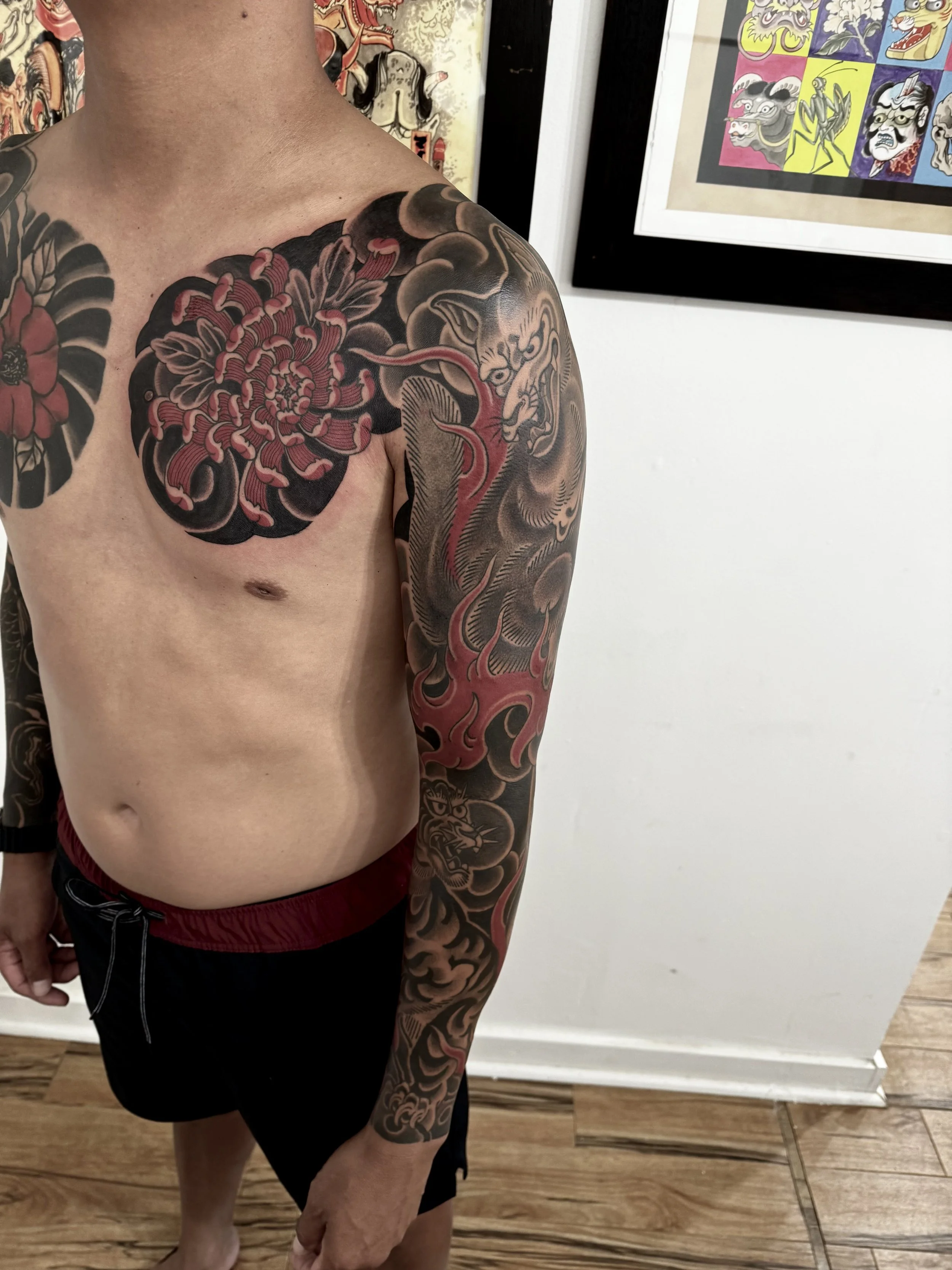 A man with tattoos on his chest, arm, and shoulder, standing in front of a framed artwork featuring traditional Japanese mask designs.
