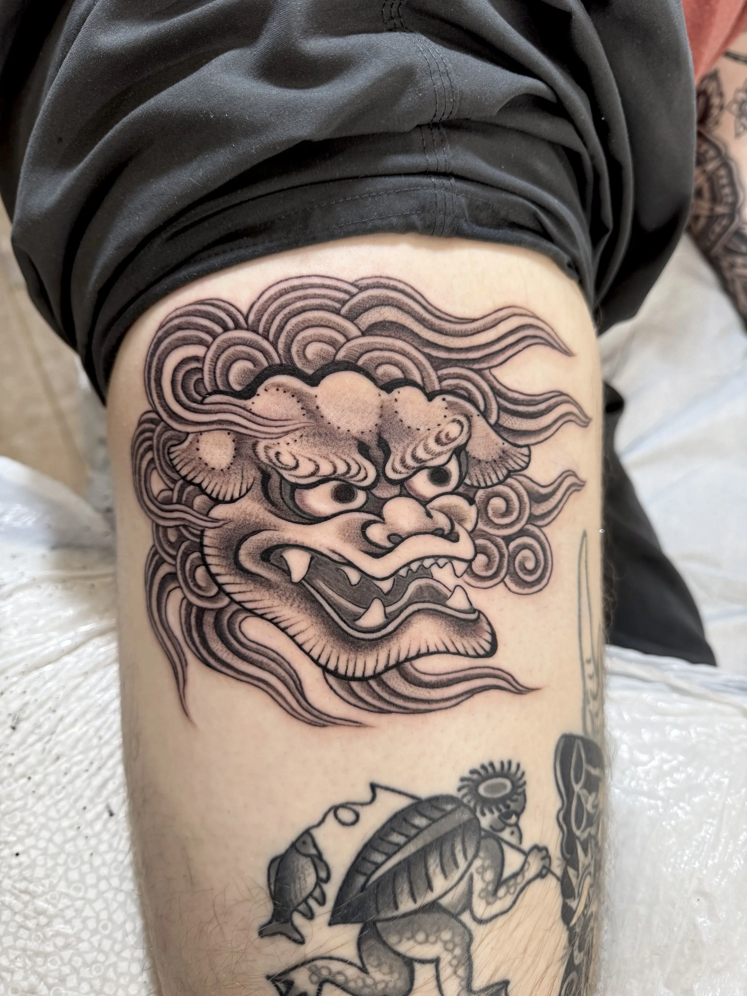 Tattoo of a fierce Chinese-style lion or dragon face with flowing mane on a person's thigh. The design is black and gray with detailed linework.