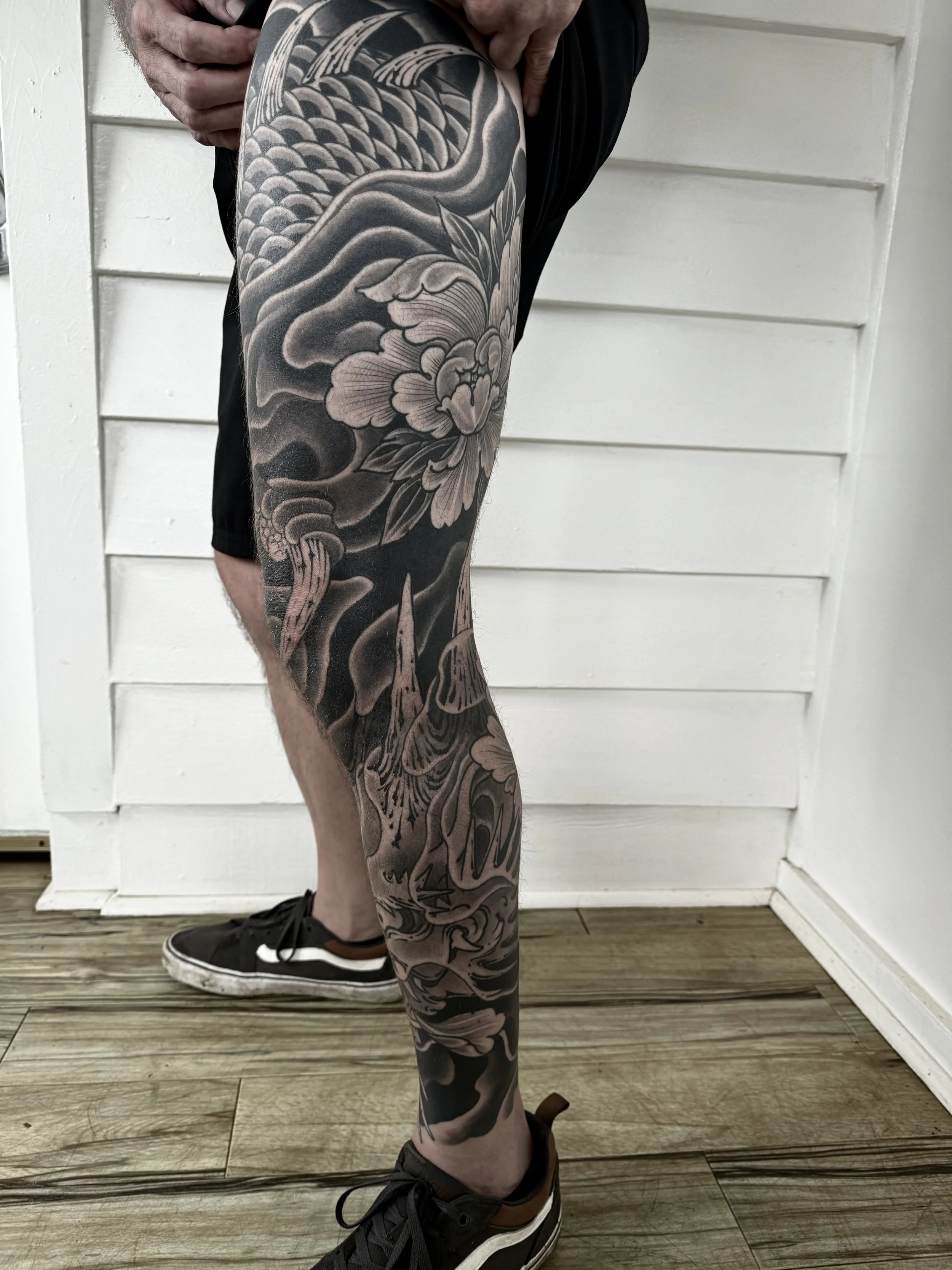 A person with a detailed black and gray Japanese-style tattoo sleeve on their left leg, featuring flowers, clouds, and mythical creatures, standing indoors near a white wooden wall, wearing black sneakers.