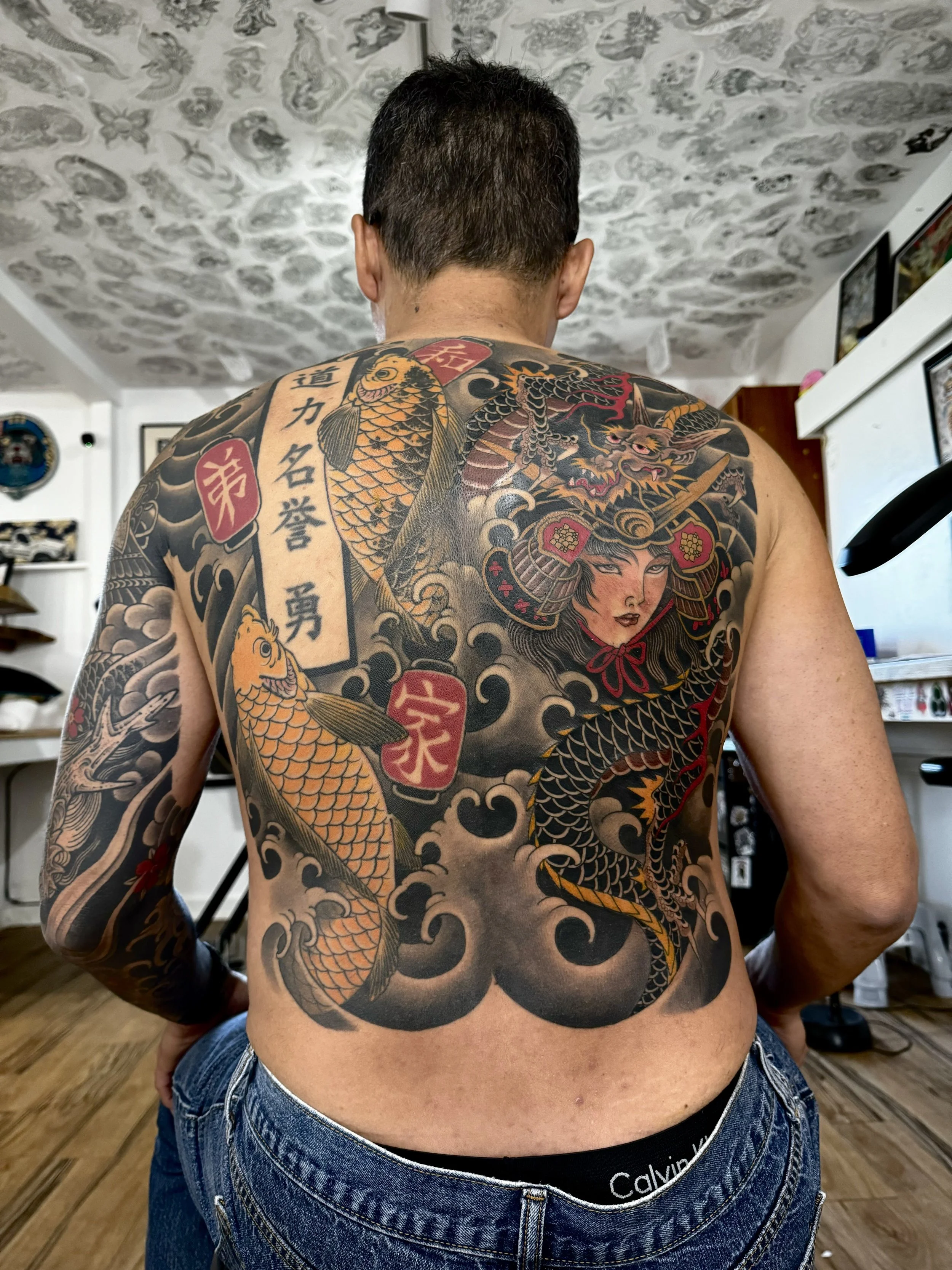 A man with a large traditional Japanese style tattoo covering his back, featuring a woman in samurai armor, a fierce dragon, two koi fish, a cloud pattern, and Japanese characters.