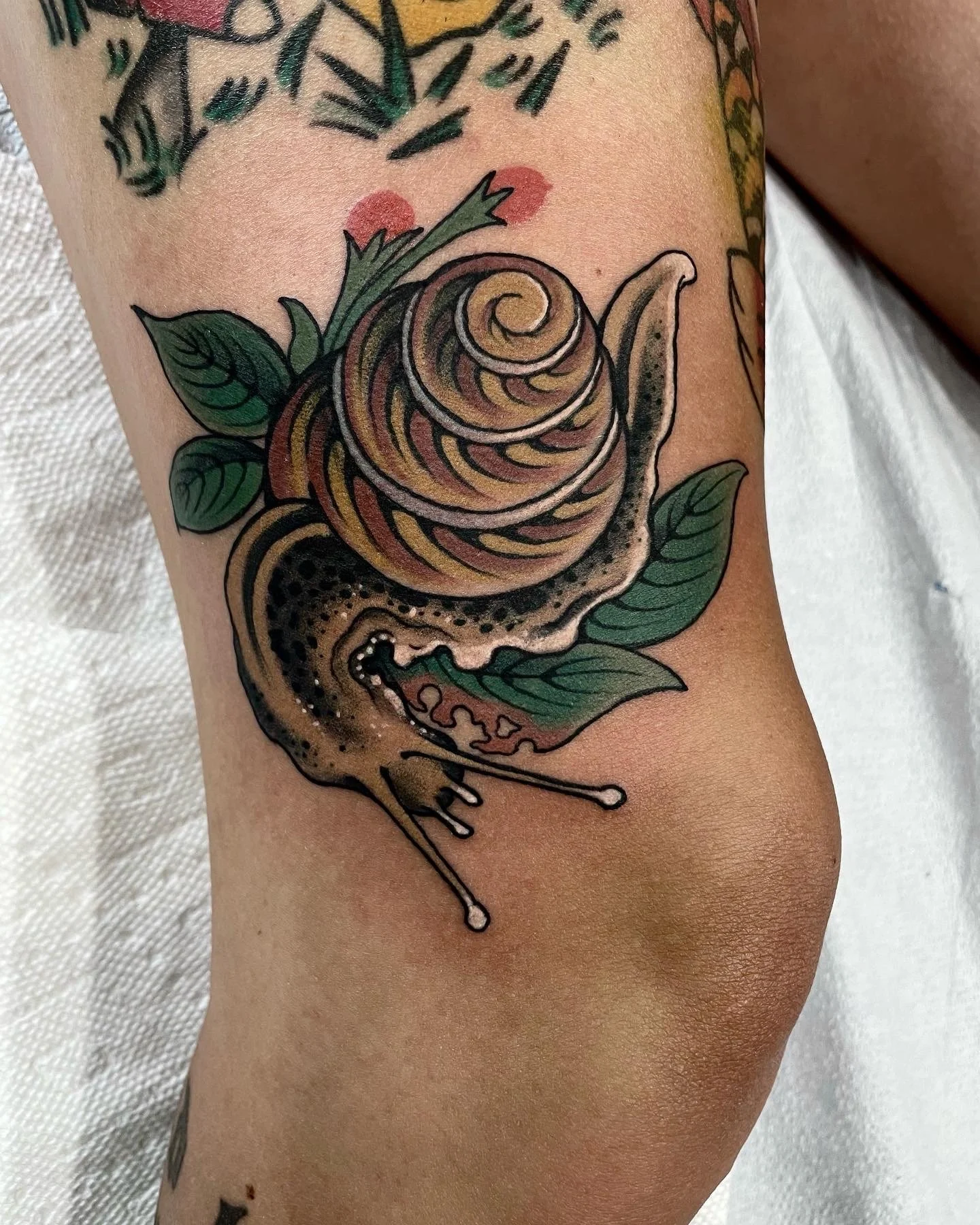 Tattoo of a snail with a coiled shell and a body, surrounded by green leaves, on a person's arm.