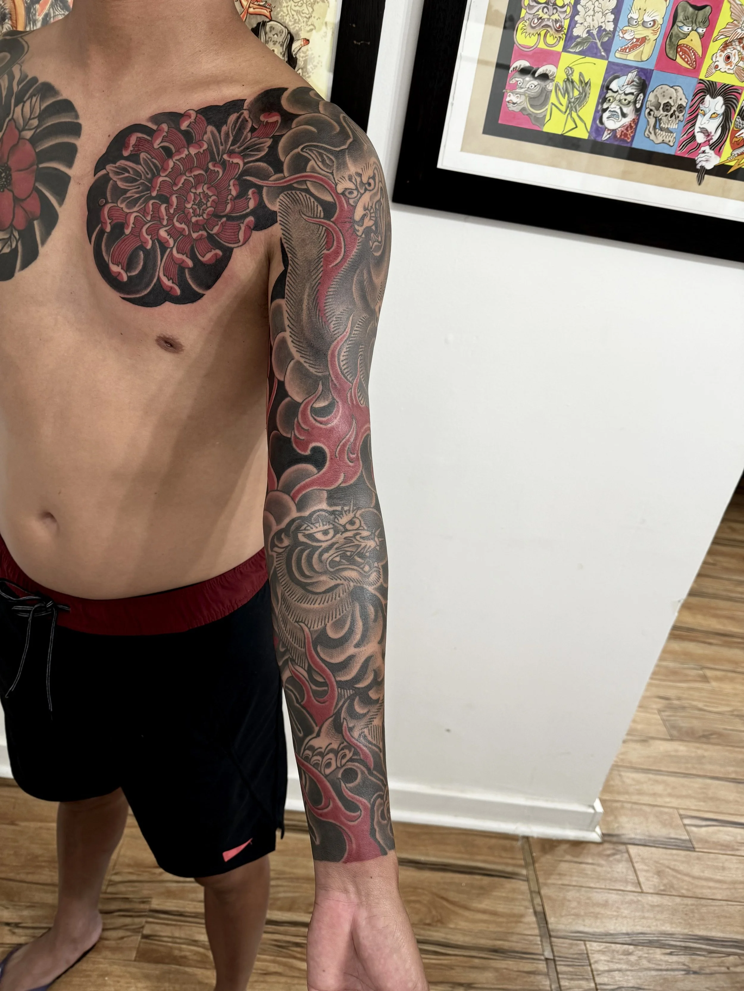 A person with a large black and red tattoo sleeve featuring a tiger, flames, and flowers on their left arm and shoulder, standing next to framed artwork of colorful, stylized horror faces on the wall.