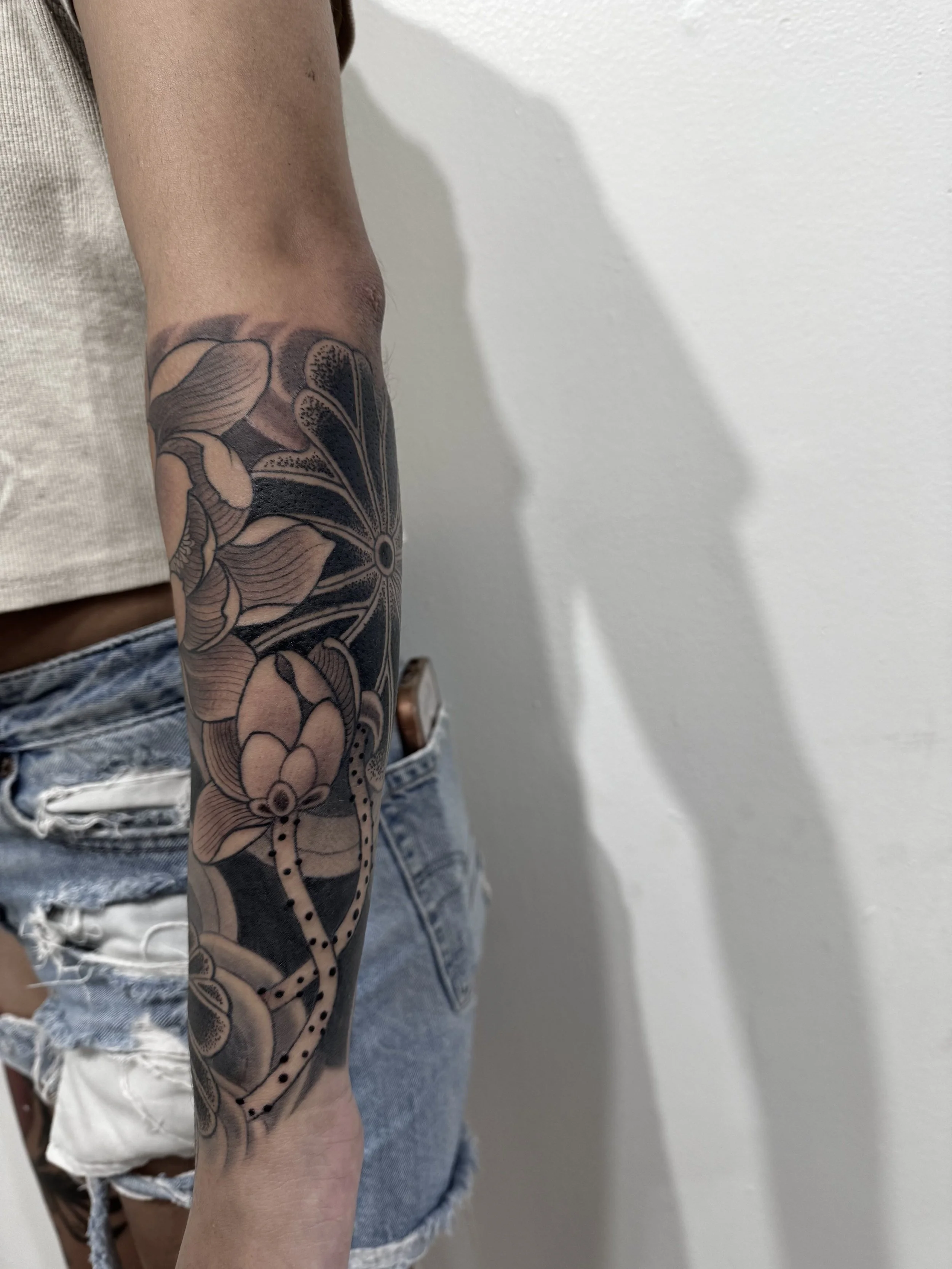A person's arm with a black and grey floral tattoo, wearing a grey sleeveless top and distressed denim shorts.