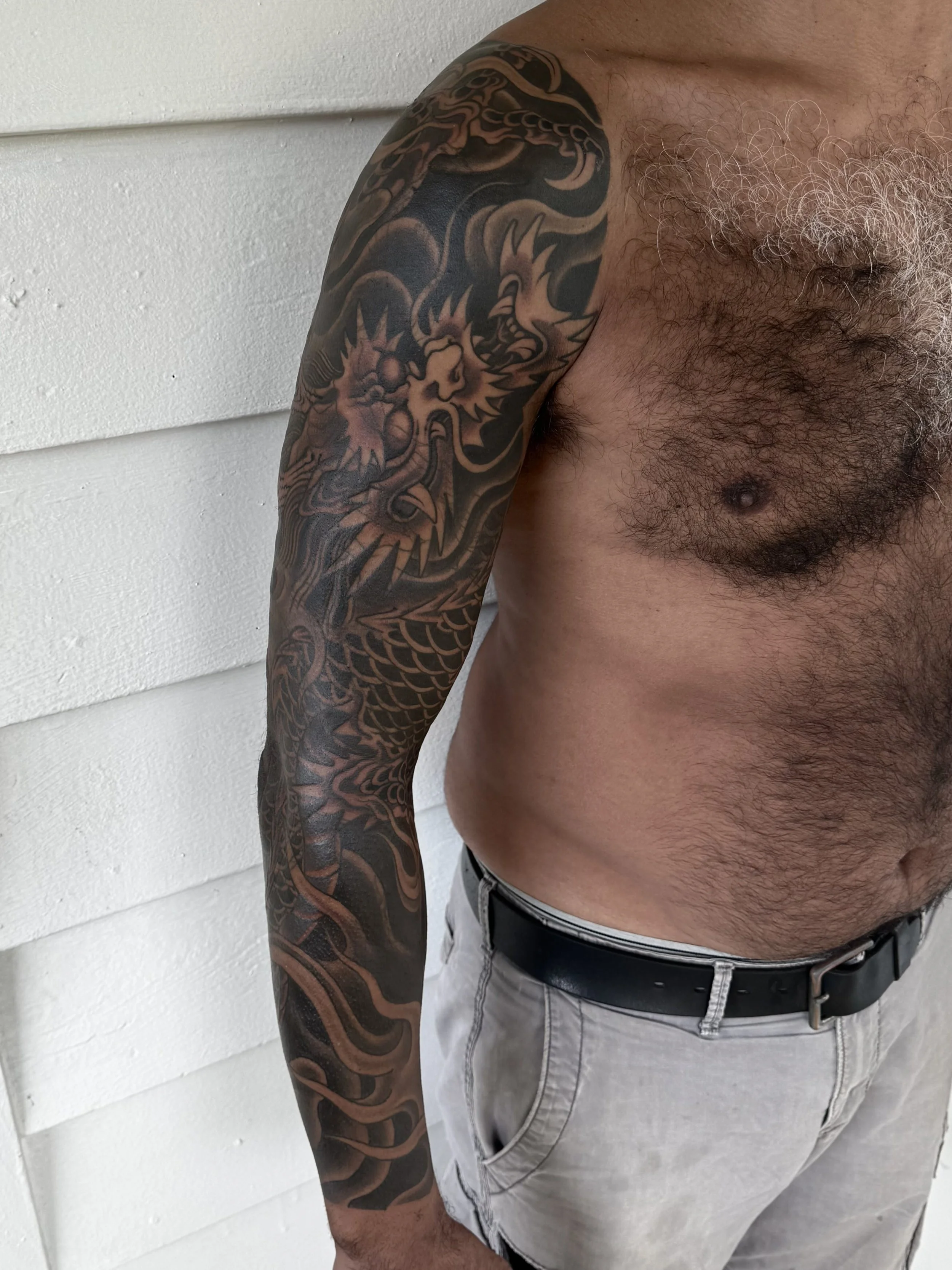 A man with a large, intricate black and gray Japanese dragon tattoo on his left arm, standing shirtless against a white wall, wearing light-colored shorts and a black belt.