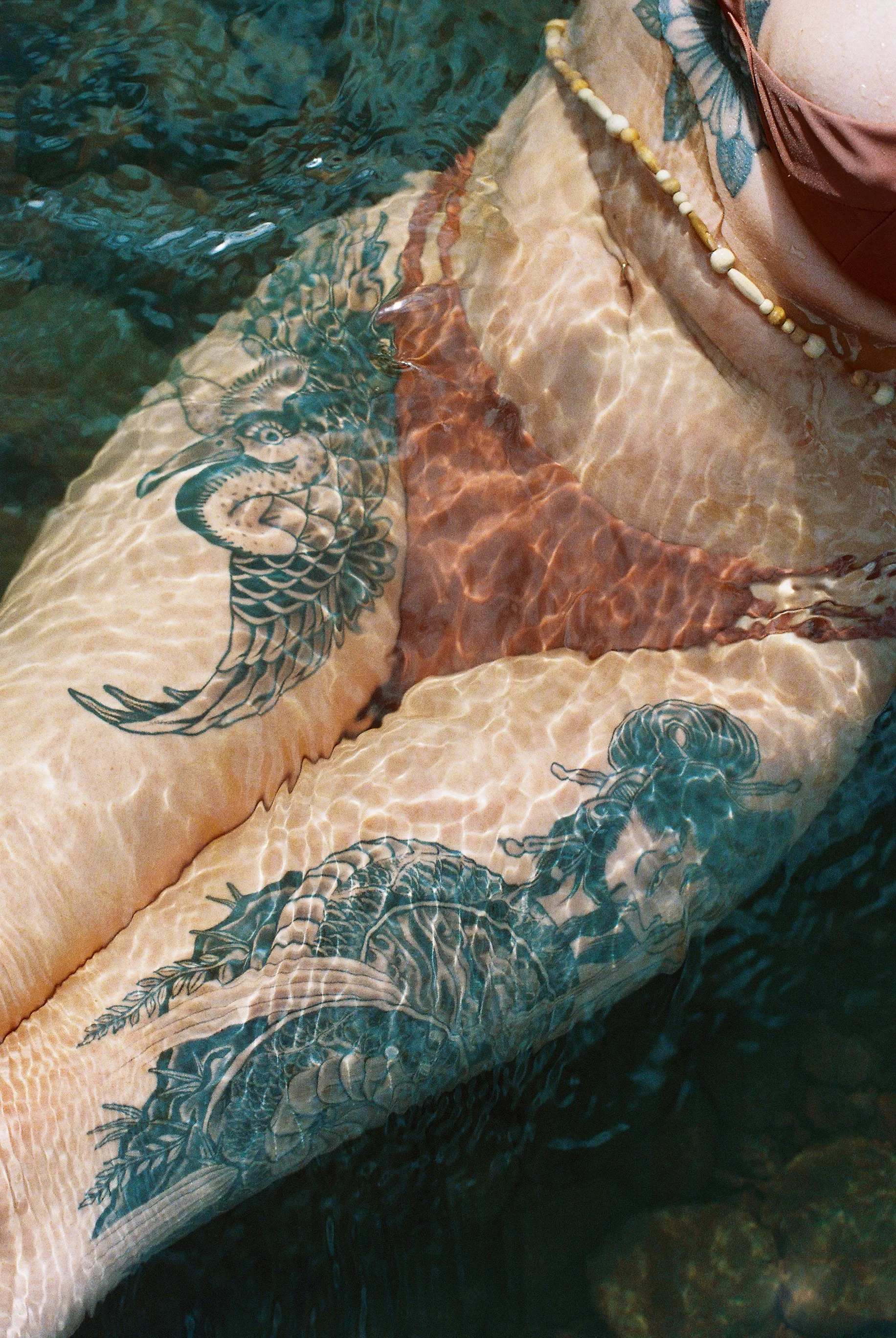 Close-up of a person's arm with tattoos under water, featuring a dragon and a butterfly, with a beaded necklace and part of a swimsuit visible.