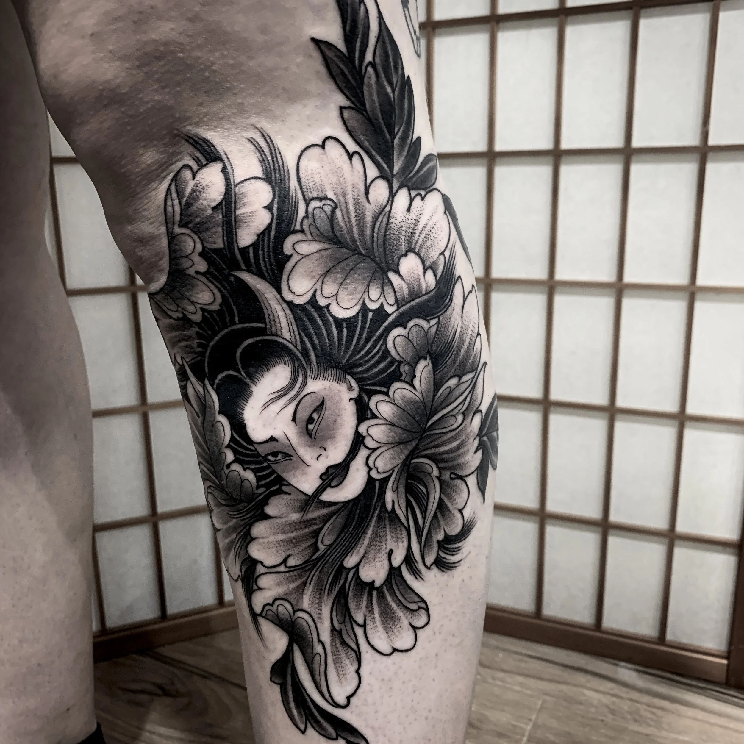 Black and gray tattoo of a woman's face surrounded by floral elements on a person's thigh.
