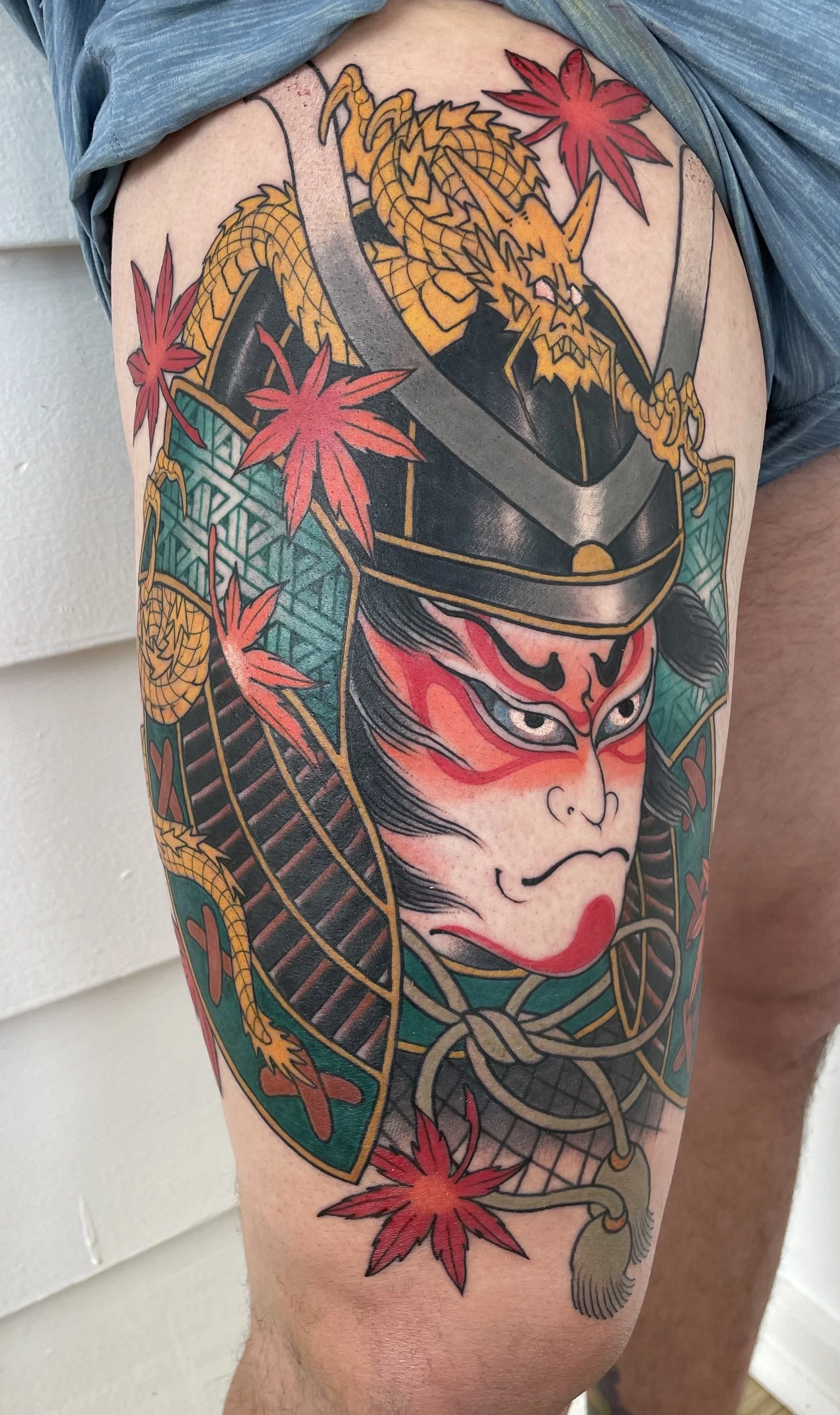 Tattoo of a samurai warrior with a fierce expression, red face paint, a helmet, and a dragon entwined with red maple leaves.