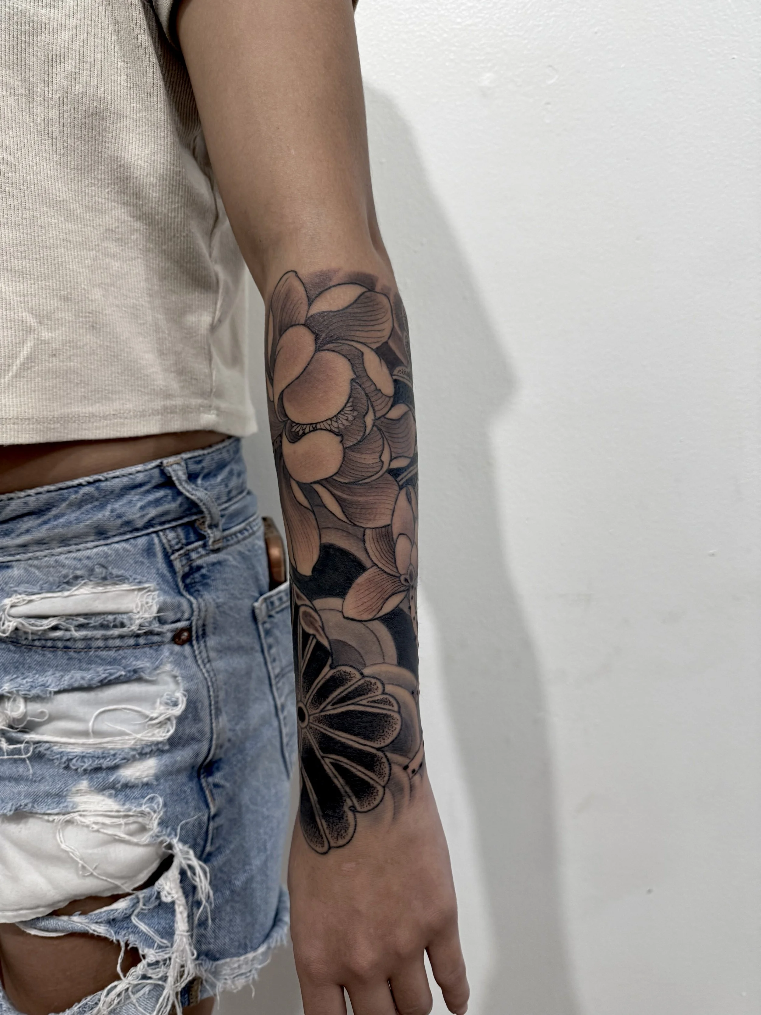 A person's right arm with a floral tattoo, featuring large flowers with detailed petals, extending from the wrist halfway up the forearm. The person is wearing a beige top and distressed denim shorts.
