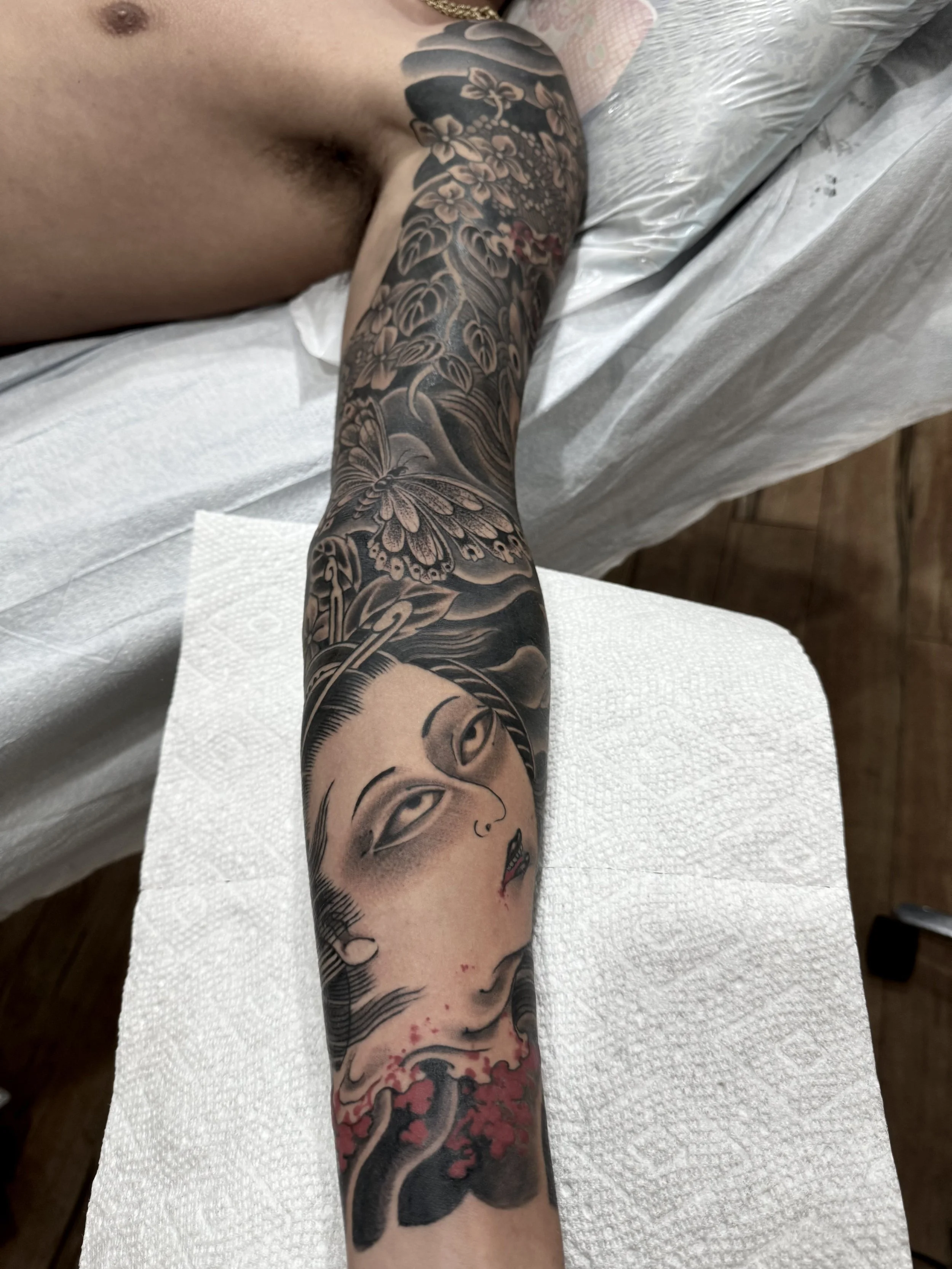 A tattoo sleeve on a person's arm featuring a woman's face with flowing hair, surrounded by various floral and butterfly designs, with some red accents near the wrist.
