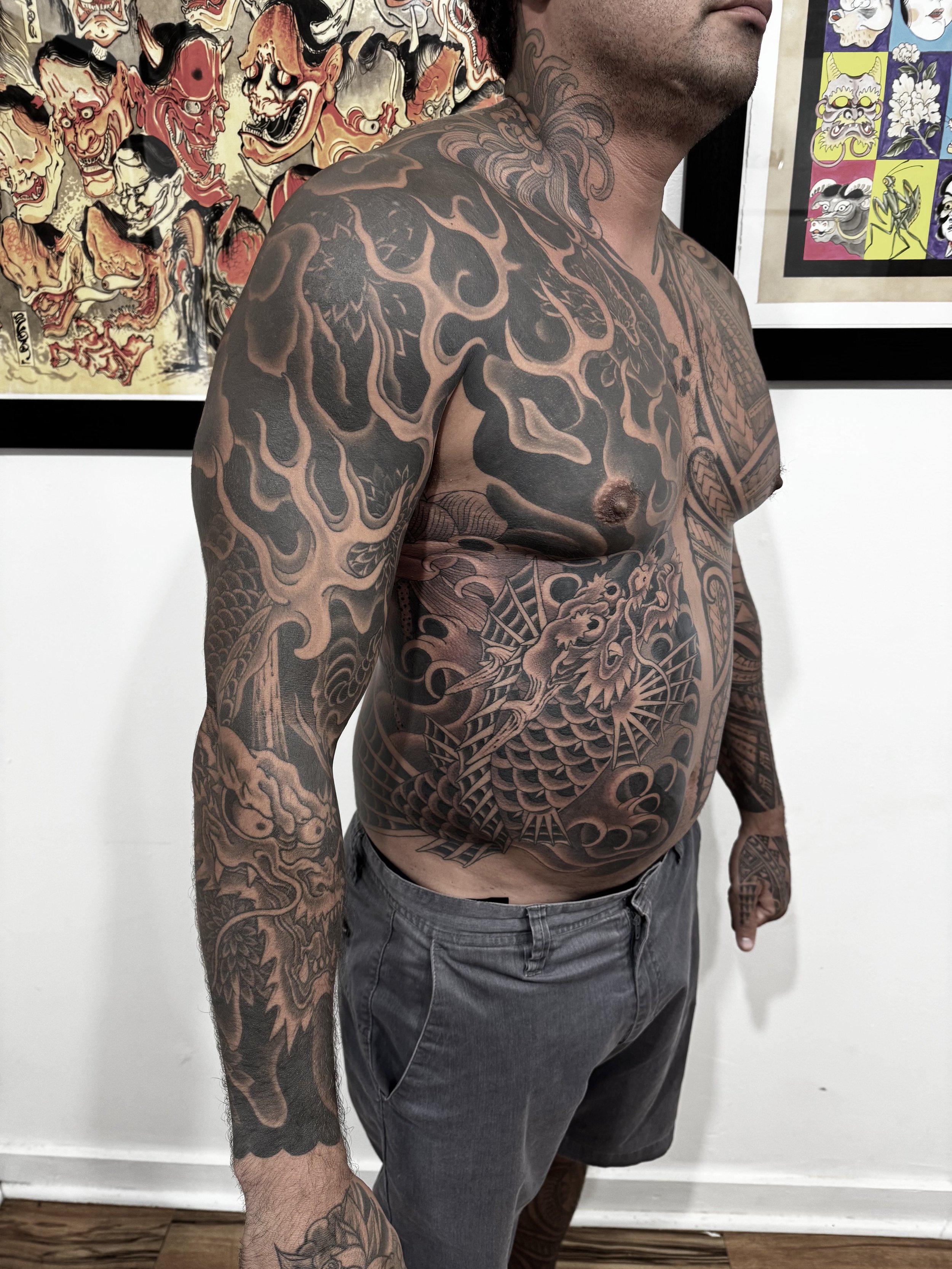 A shirtless man with extensive black and gray traditional Japanese style tattoos covering his upper body, including his shoulders, chest, and arms. The tattoos feature dragons, waves, and other mythic imagery. He is standing in an art gallery with co