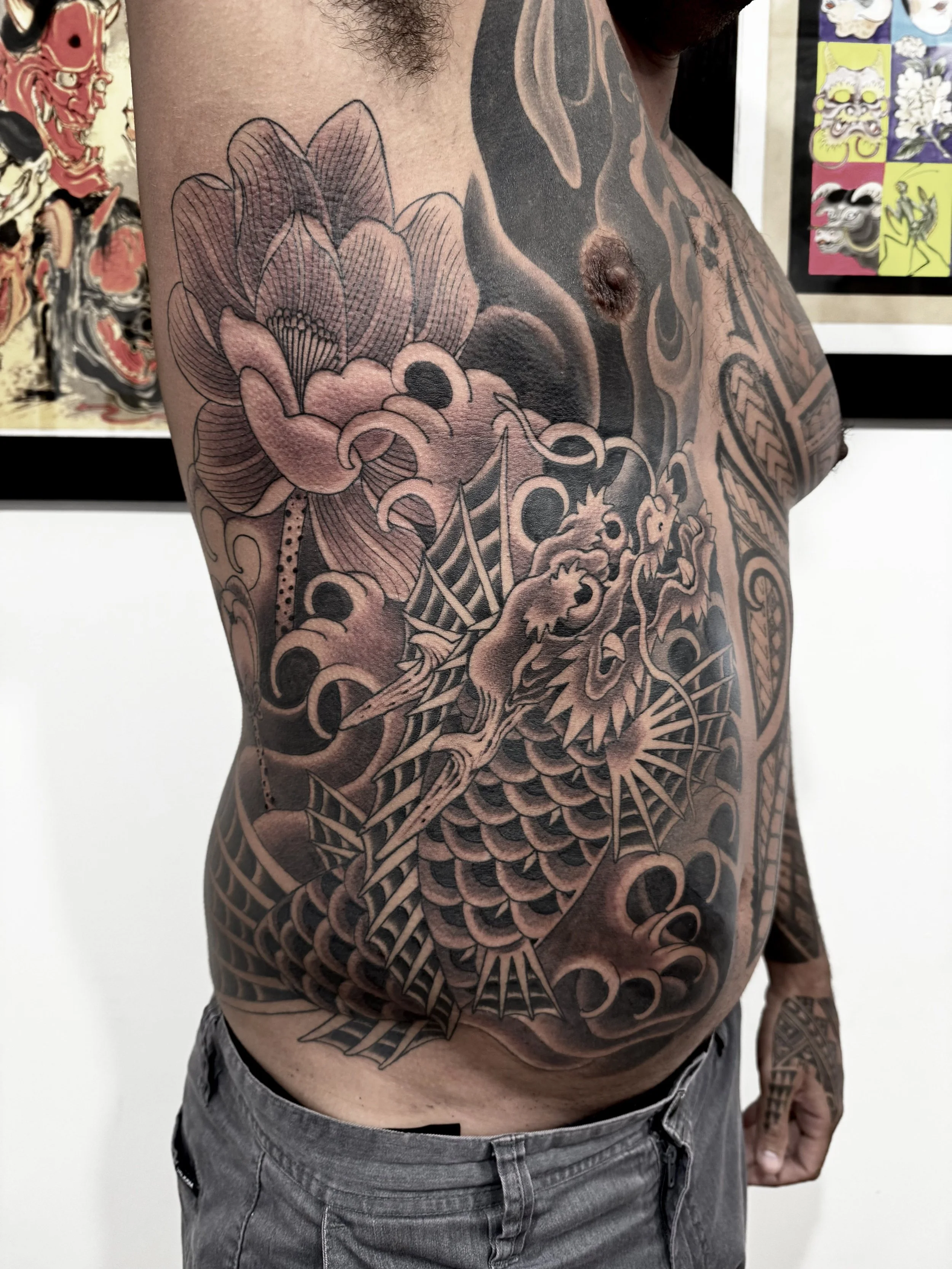 A person with extensive black and gray tattoos covering the left side of their chest and torso, featuring a detailed dragon wrapped around a stylized flower and geometric patterns.