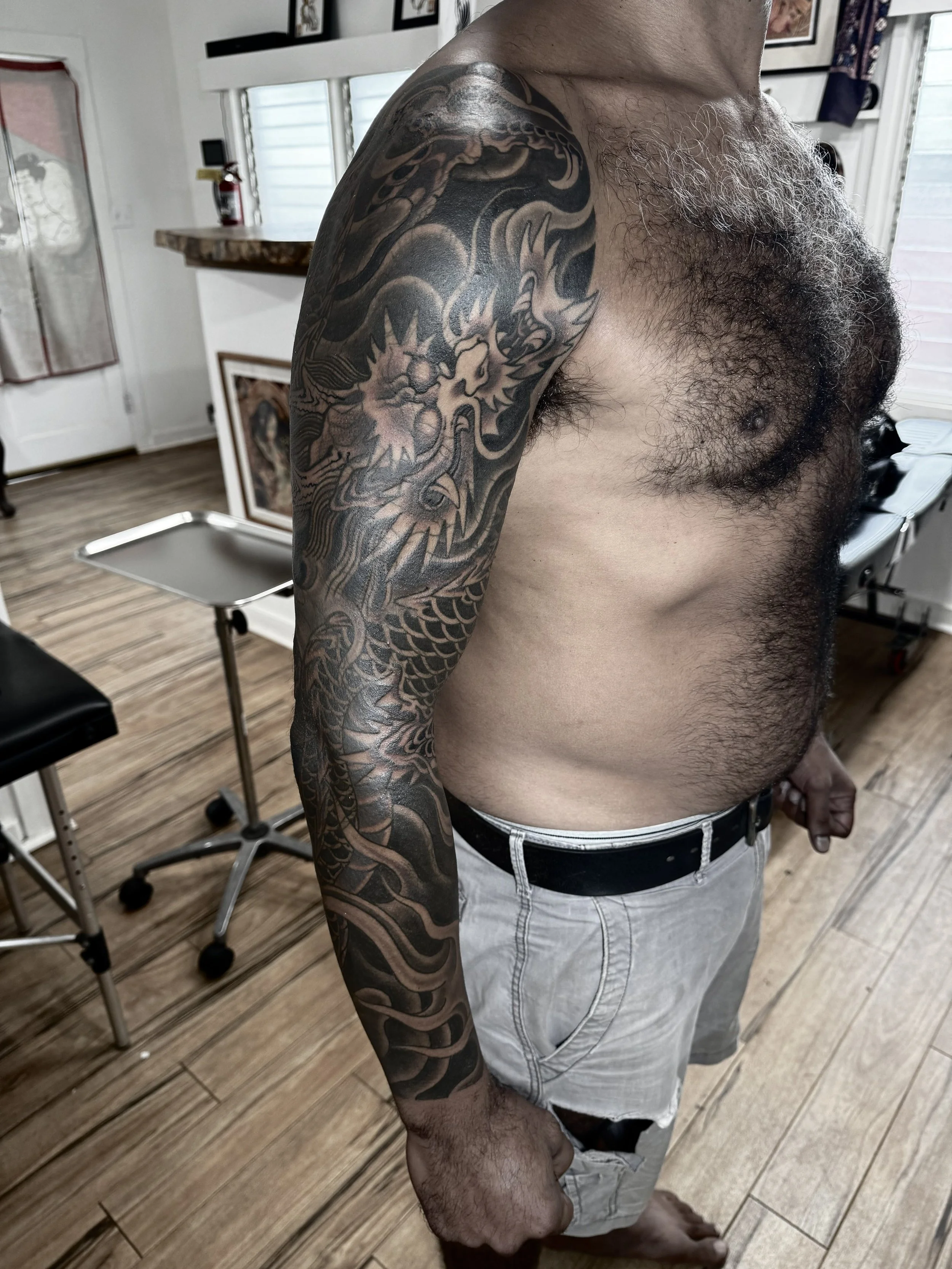 A man with a detailed black and gray dragon tattoo covering his upper arm and shoulder, standing in a room with wooden flooring and various furniture.