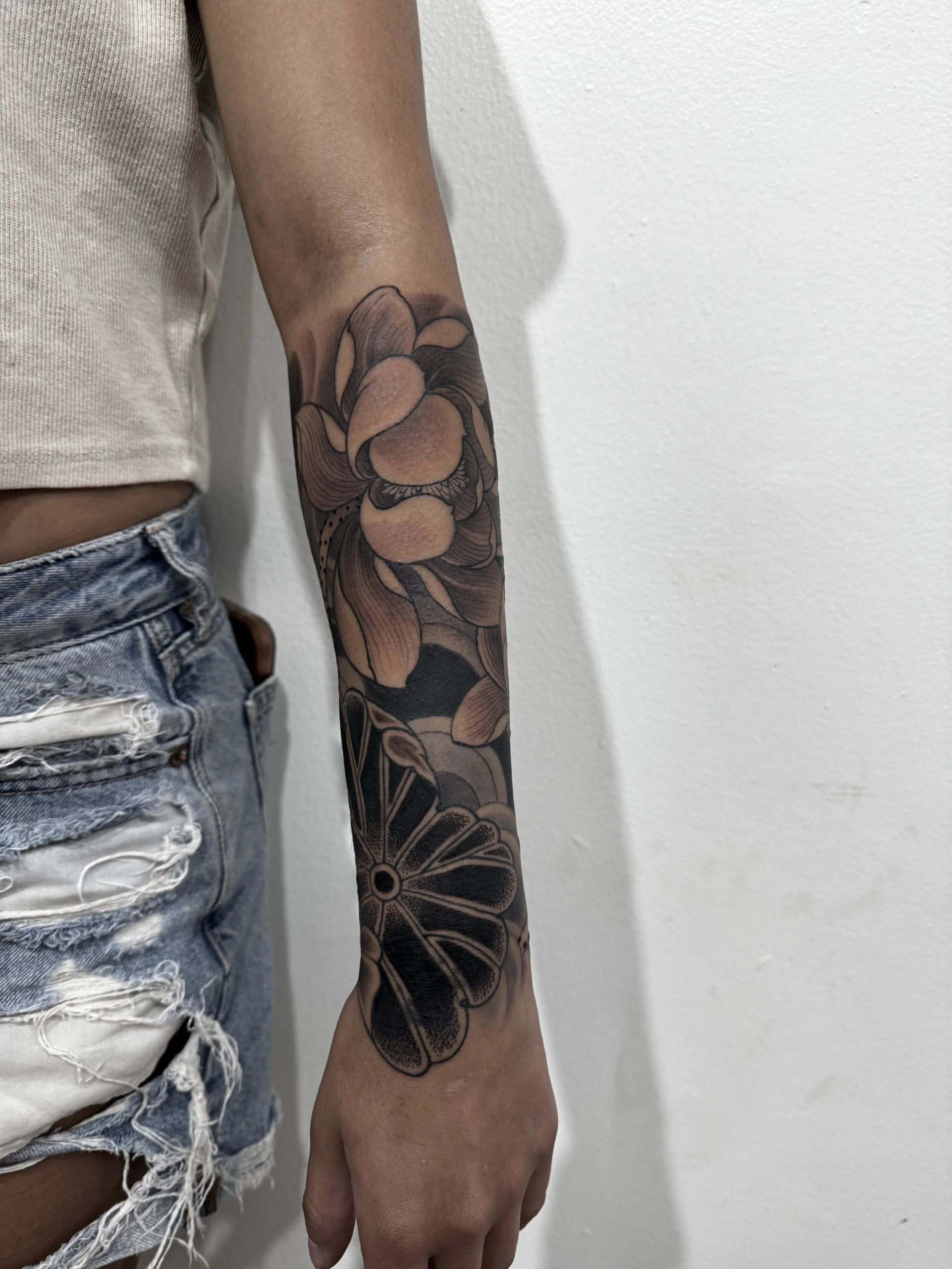 Tattoo of a flower and a citrus slice on a person's forearm, with casual ripped denim shorts and a beige top.